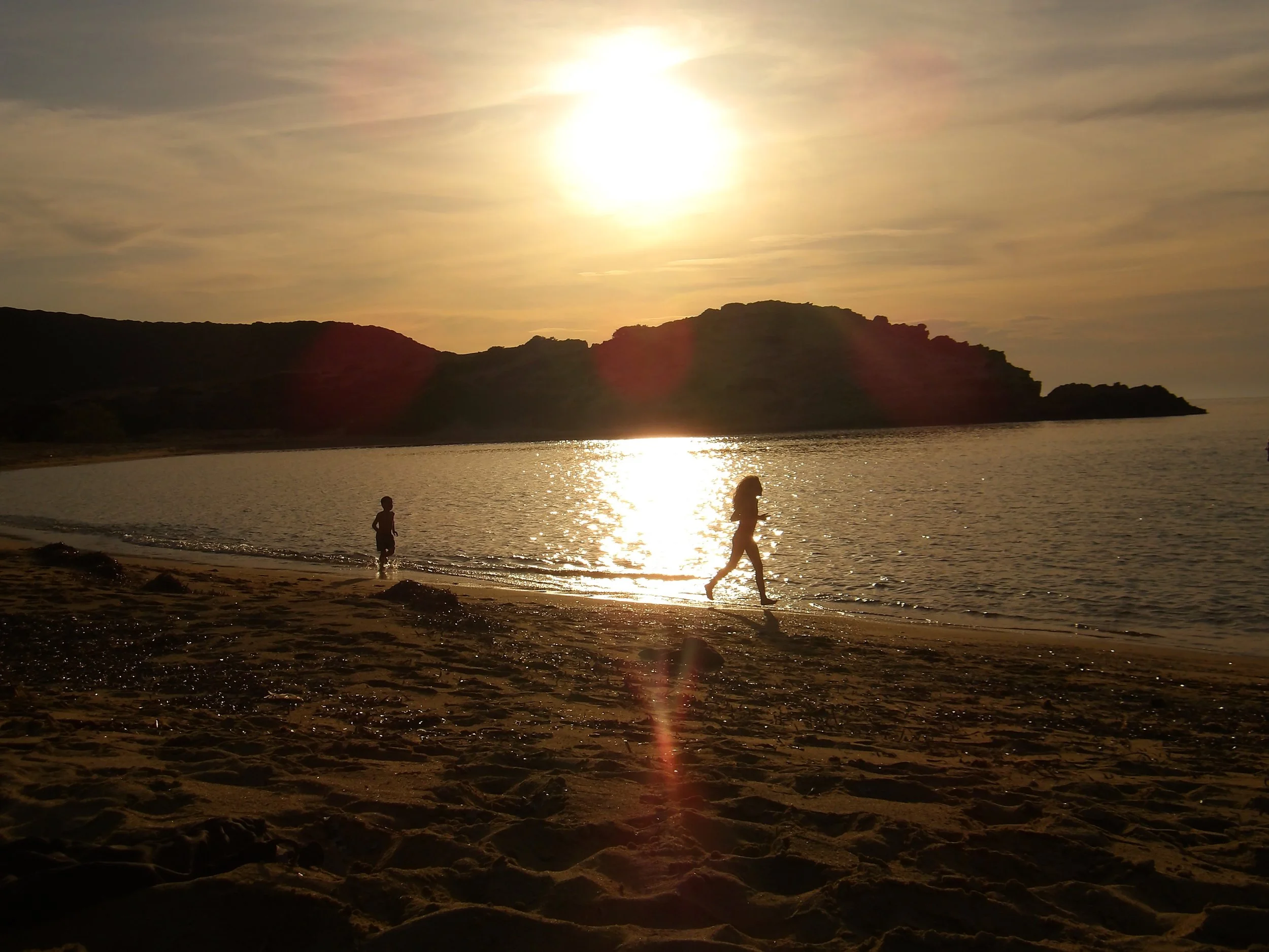 Sunset over a beach with two children playing in the shallow water, rocky formations in the foreground, and a hill or cliff in the background.
