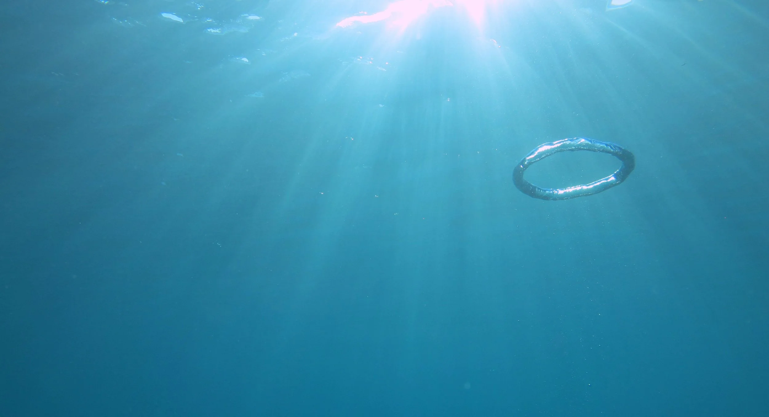 An underwater scene with sunlight filtering through the water and a small circular object, possibly a ring, floating in the water.