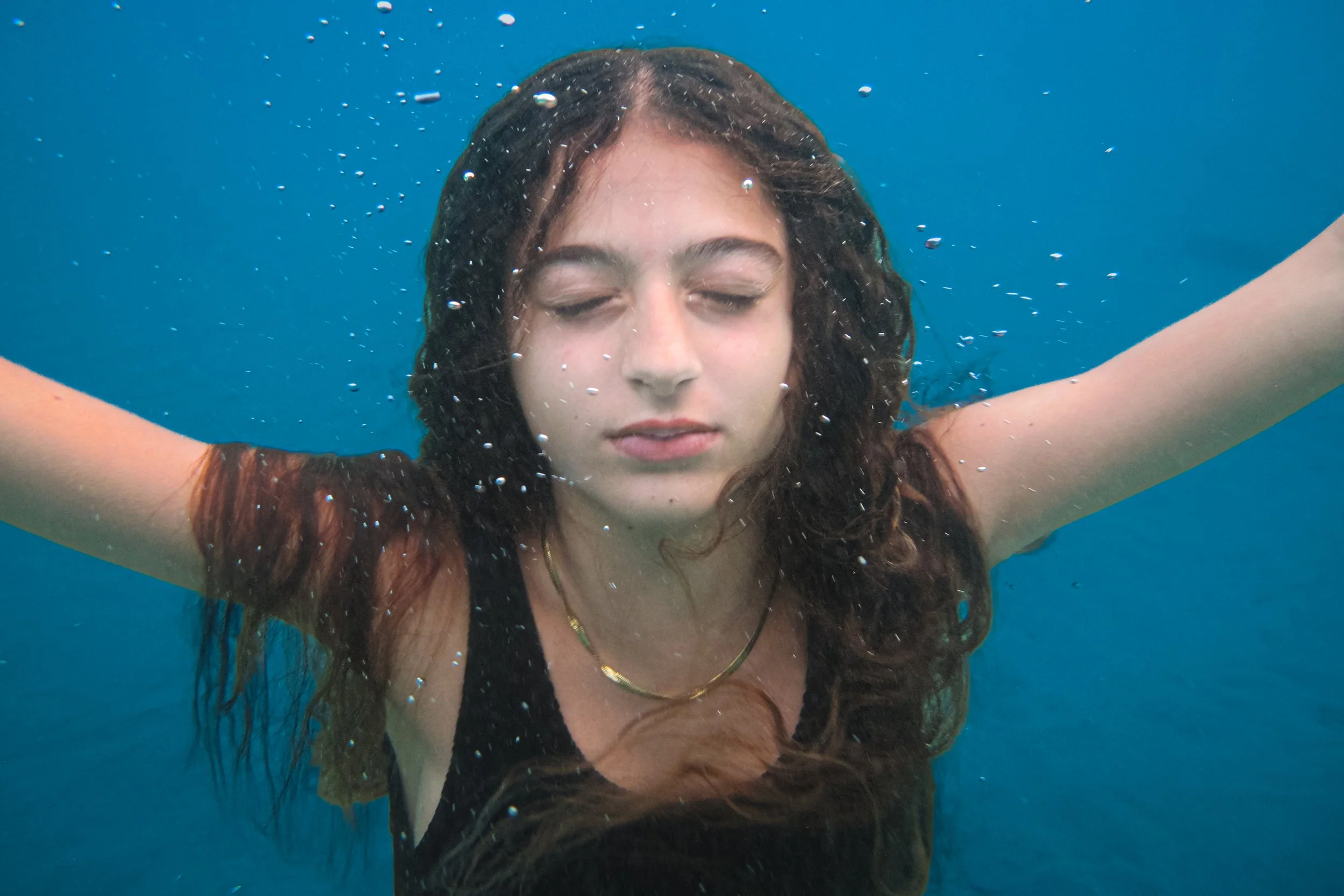 A woman with long brown hair floating underwater with her eyes closed.