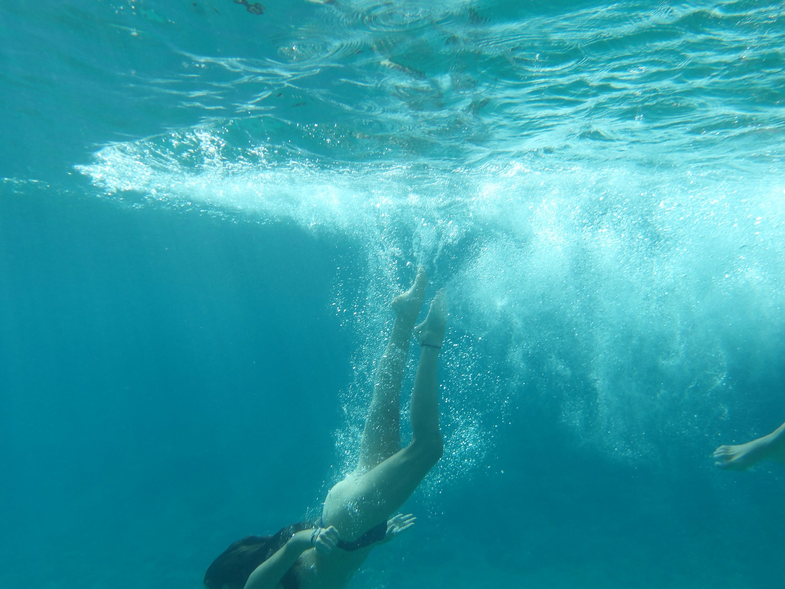 A person swimming underwater in a pool with arms extended forward and hair floating, creating bubbles and ripples in the water.