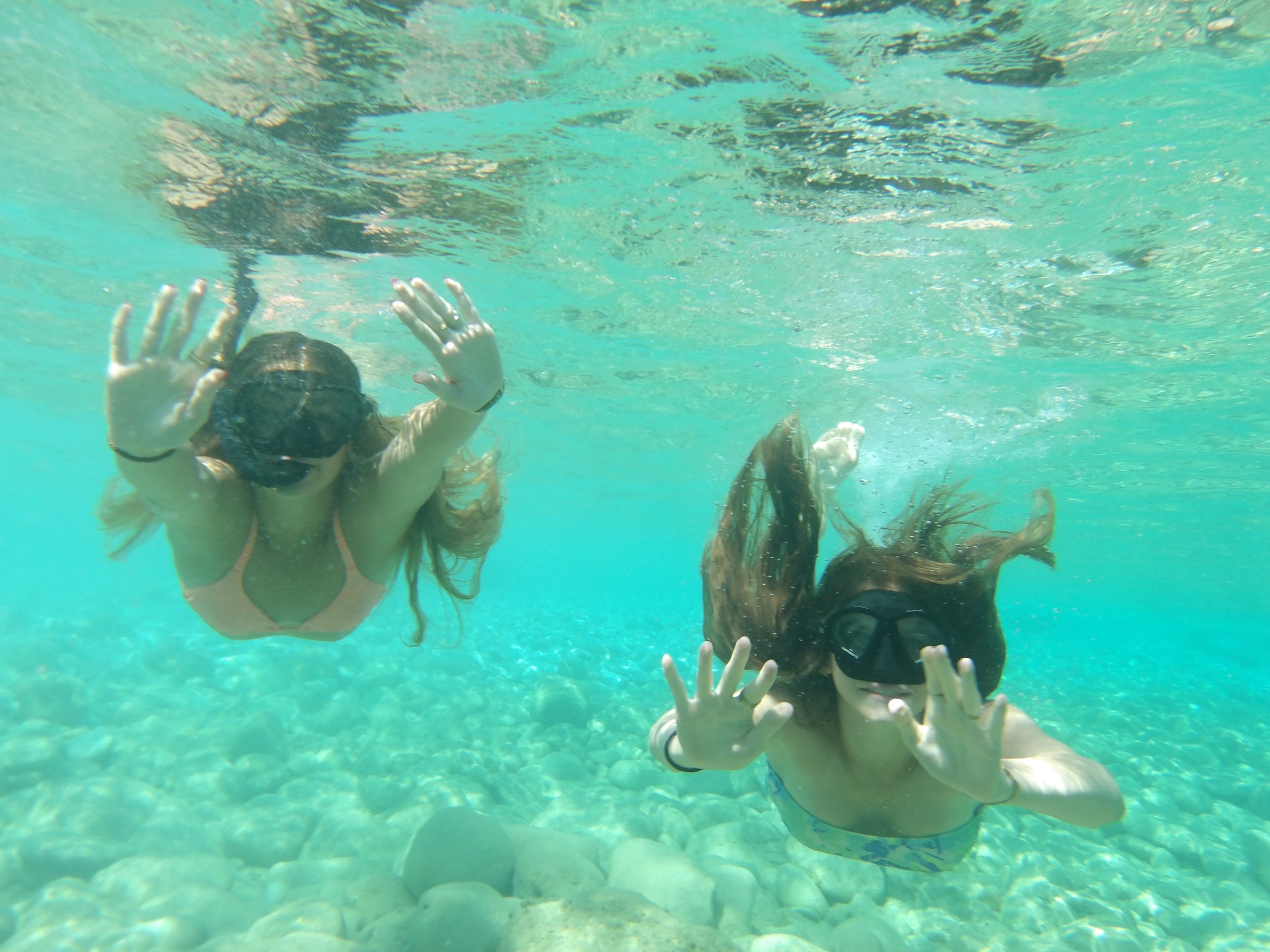 Two children swimming underwater in a clear pool, reaching towards the camera with their hands, with bubbles and sunlight visible on the water's surface.