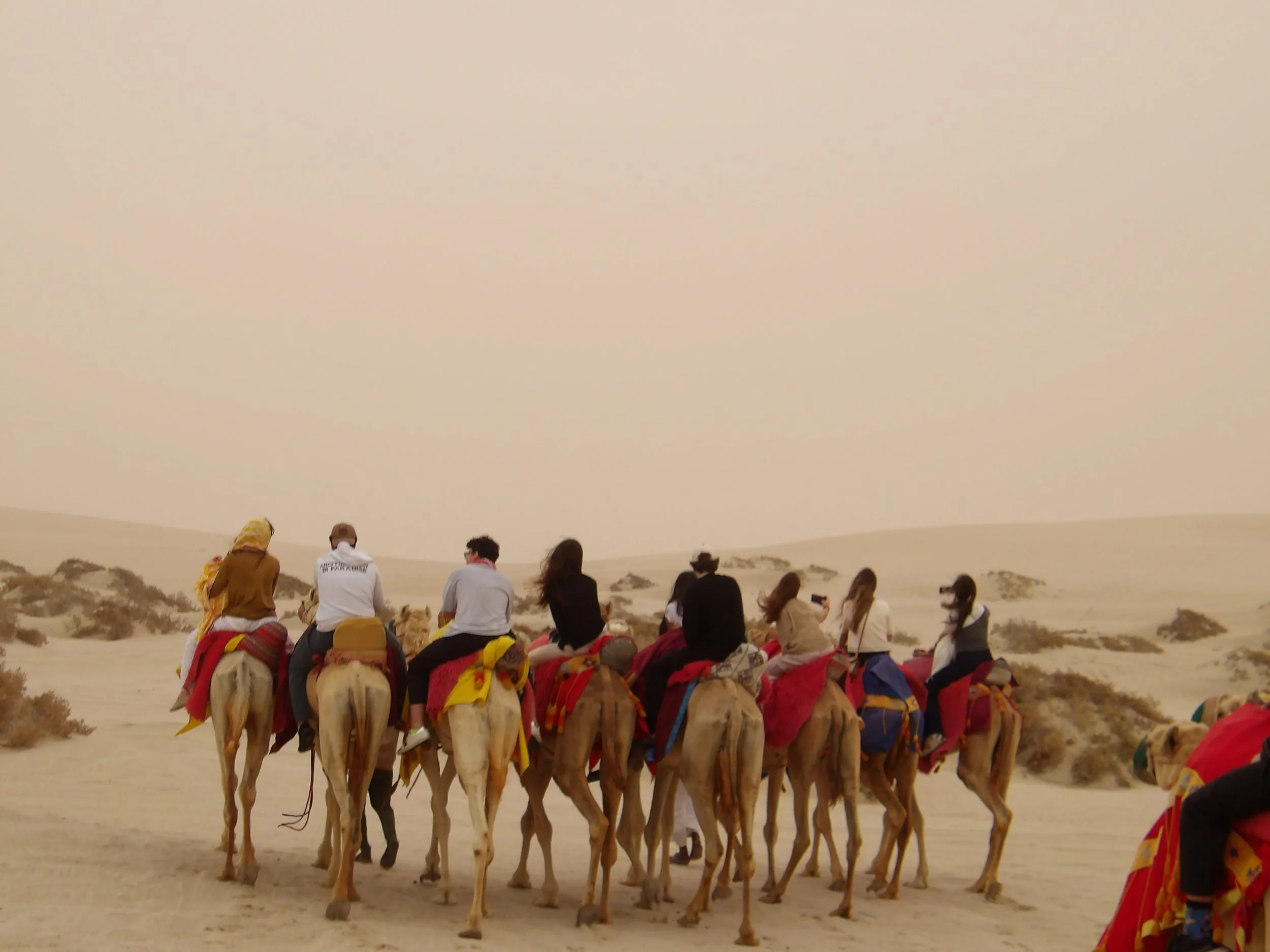 Riding camels in a desert landscape under a hazy sky, with some people taking photos and wearing hats and scarves.