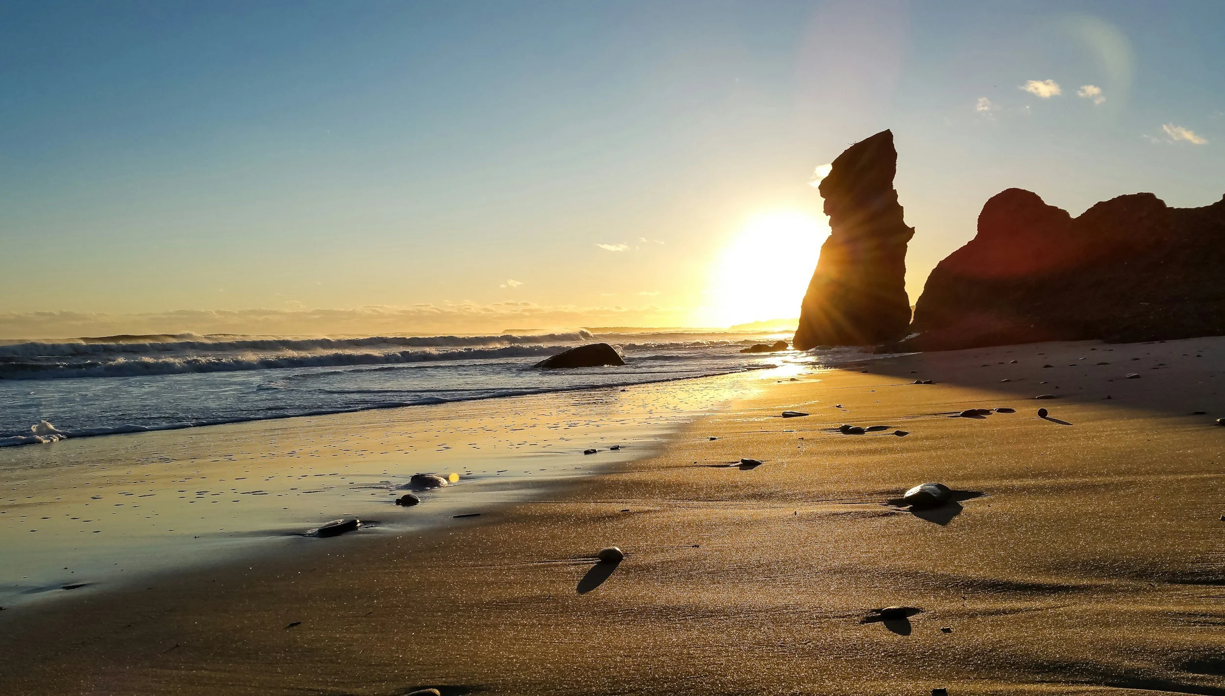 A beach scene at sunset with large rock formations near the shoreline, waves crashing, and a clear sky with a few clouds.