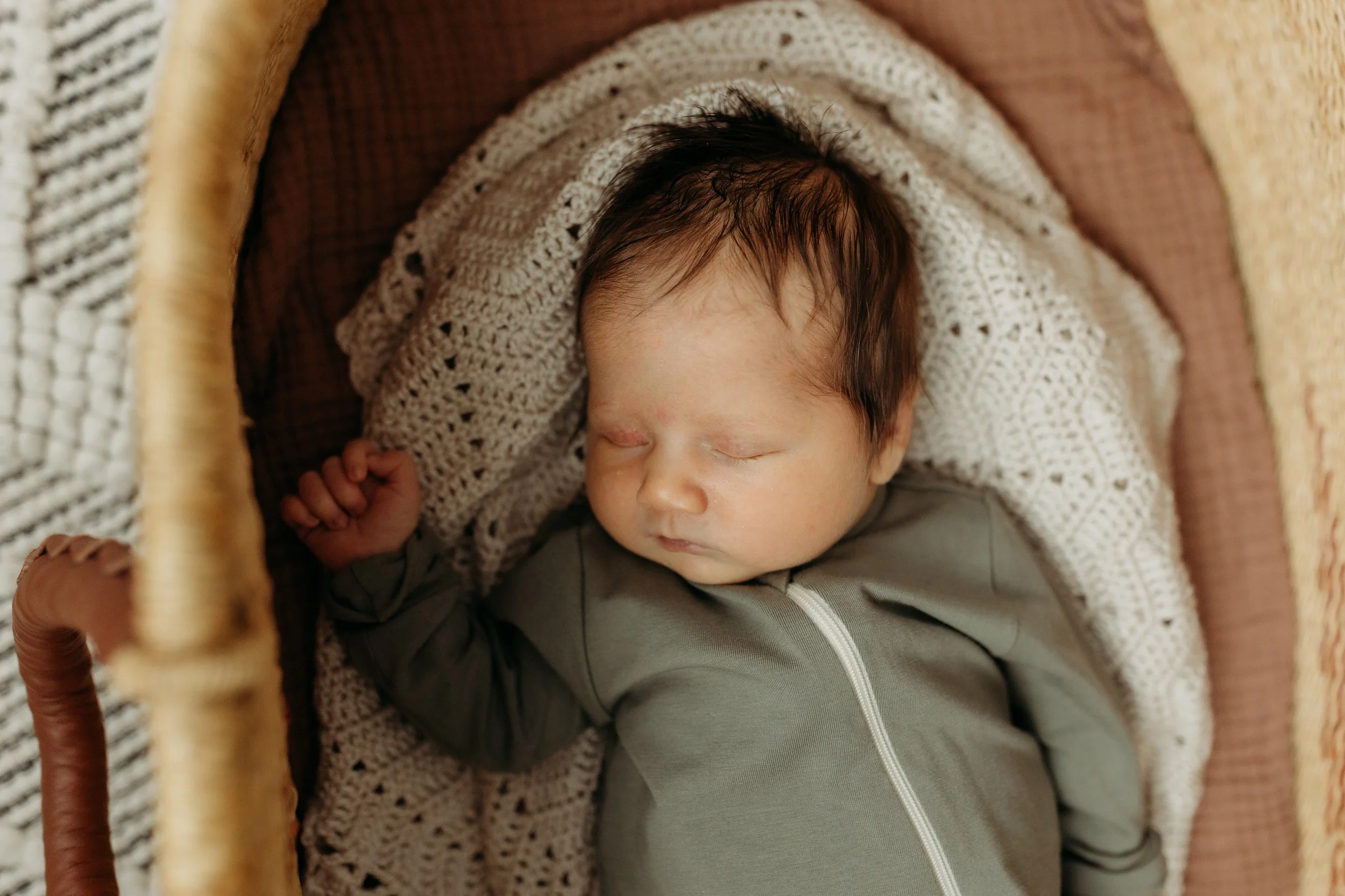 A sleeping baby with dark hair tucked in a knit blanket inside a woven bassinet.