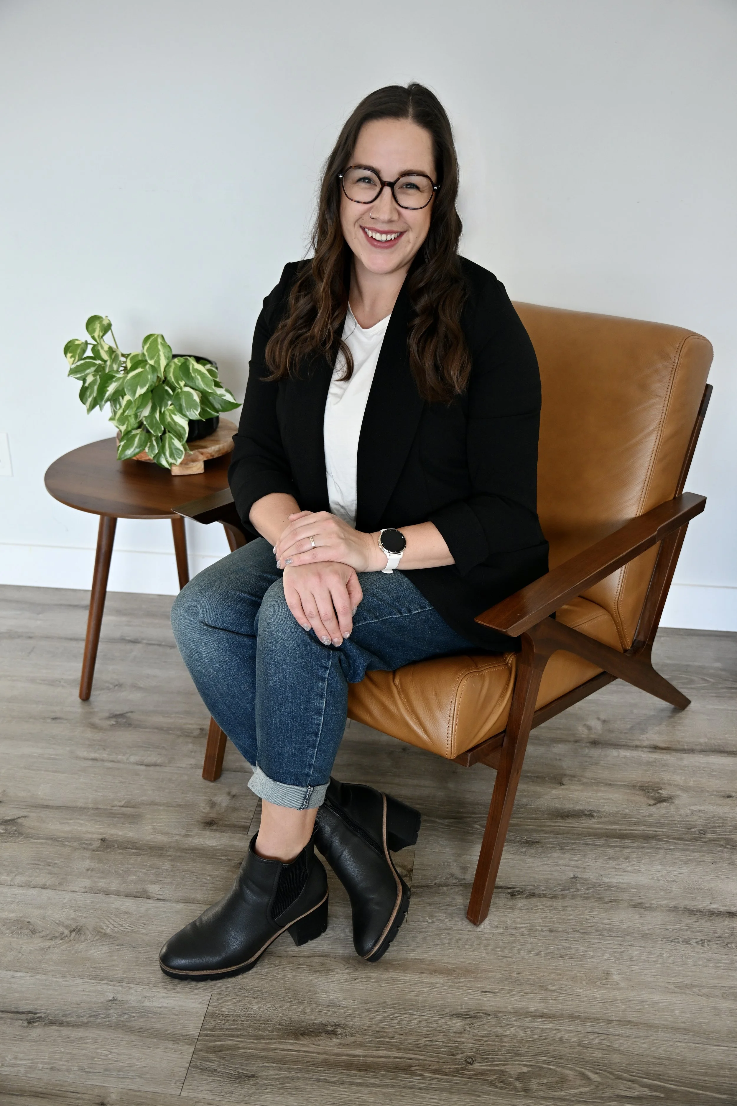 A woman with long brown hair, glasses, and a white shirt under a black blazer sitting on a brown leather armchair. She is smiling, with her hands clasped on her lap. A small wooden side table with a potted green plant is behind her, and she is wearing dark jeans and black ankle boots. The room has a light-colored wall and wood-look flooring.
