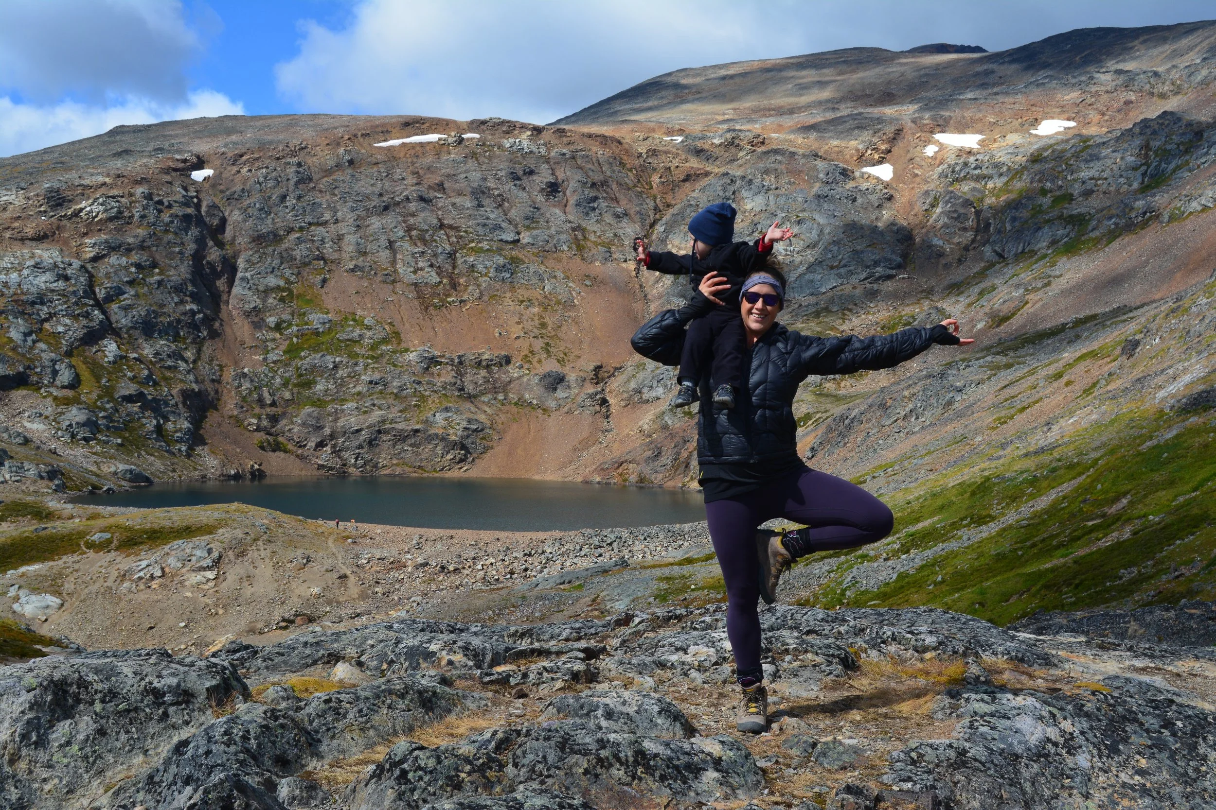 A woman practicing yoga in a tree pose with one leg raised and hands in a mudra, standing on rocky terrain near a mountain lake, with a child sitting on her shoulders, both smiling. The landscape features rugged mountains, some patches of snow, and a partly cloudy sky.