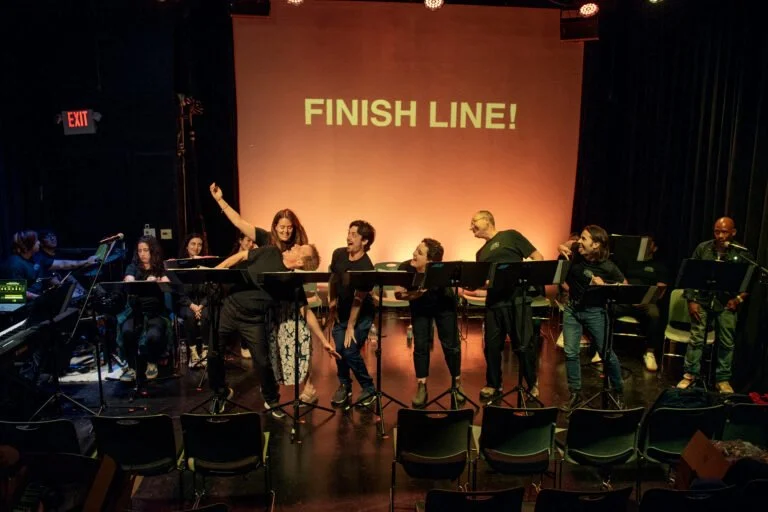 Group of musicians performing on stage with a large screen behind them displaying 'Finish Line!' in yellow letters. The performers include a keyboardist on the left, a conductor, and several other musicians with instruments, under stage lighting.