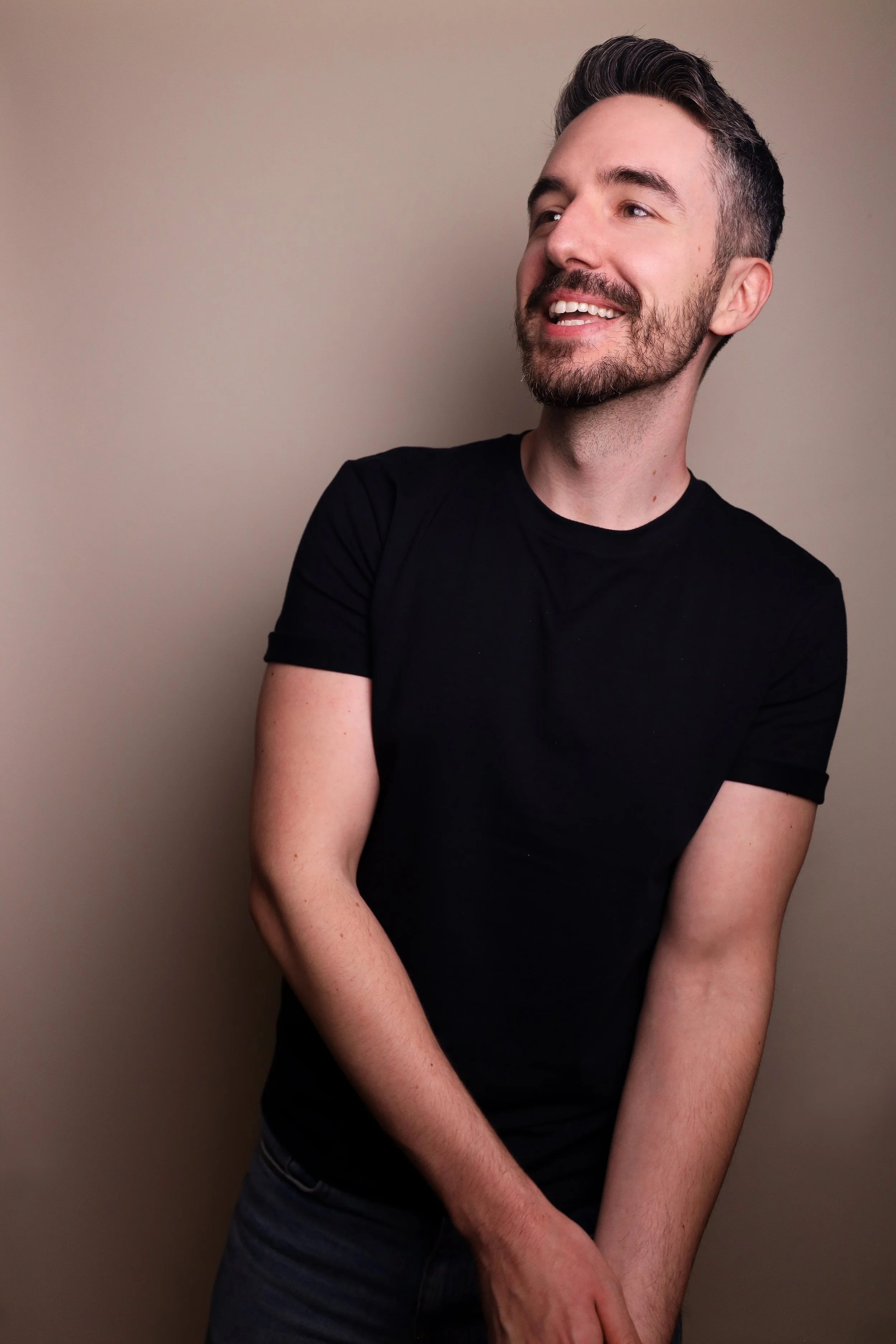 A young man with short, dark, styled hair and a beard, wearing a black t-shirt, smiling and looking to the side against a plain, neutral-colored background.