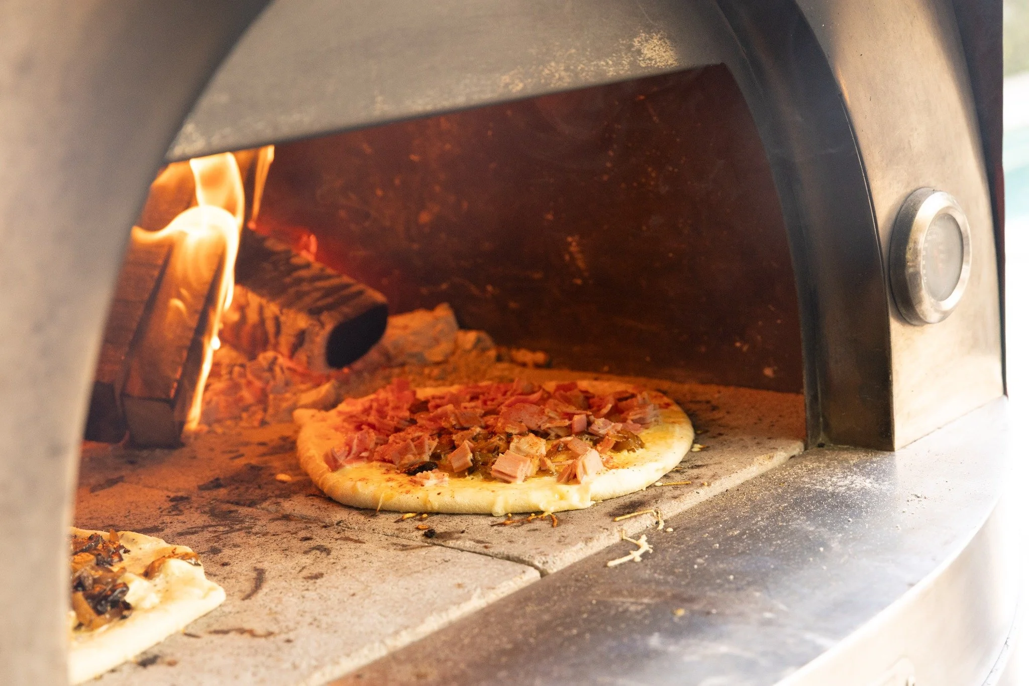 Pizza baking beside flames in a wood-fired oven.