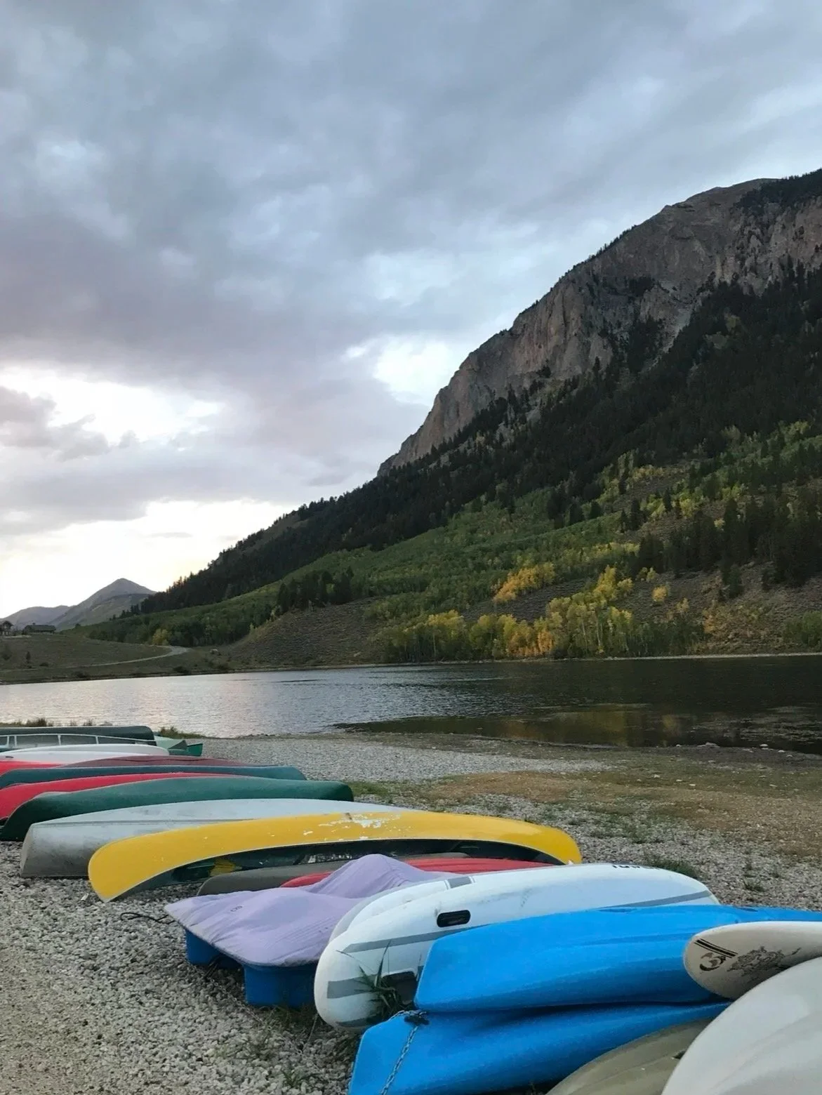 Colorful kayaks and paddleboards lined up on a rocky shore beside a calm lake surrounded by green trees and mountains under cloudy sky.