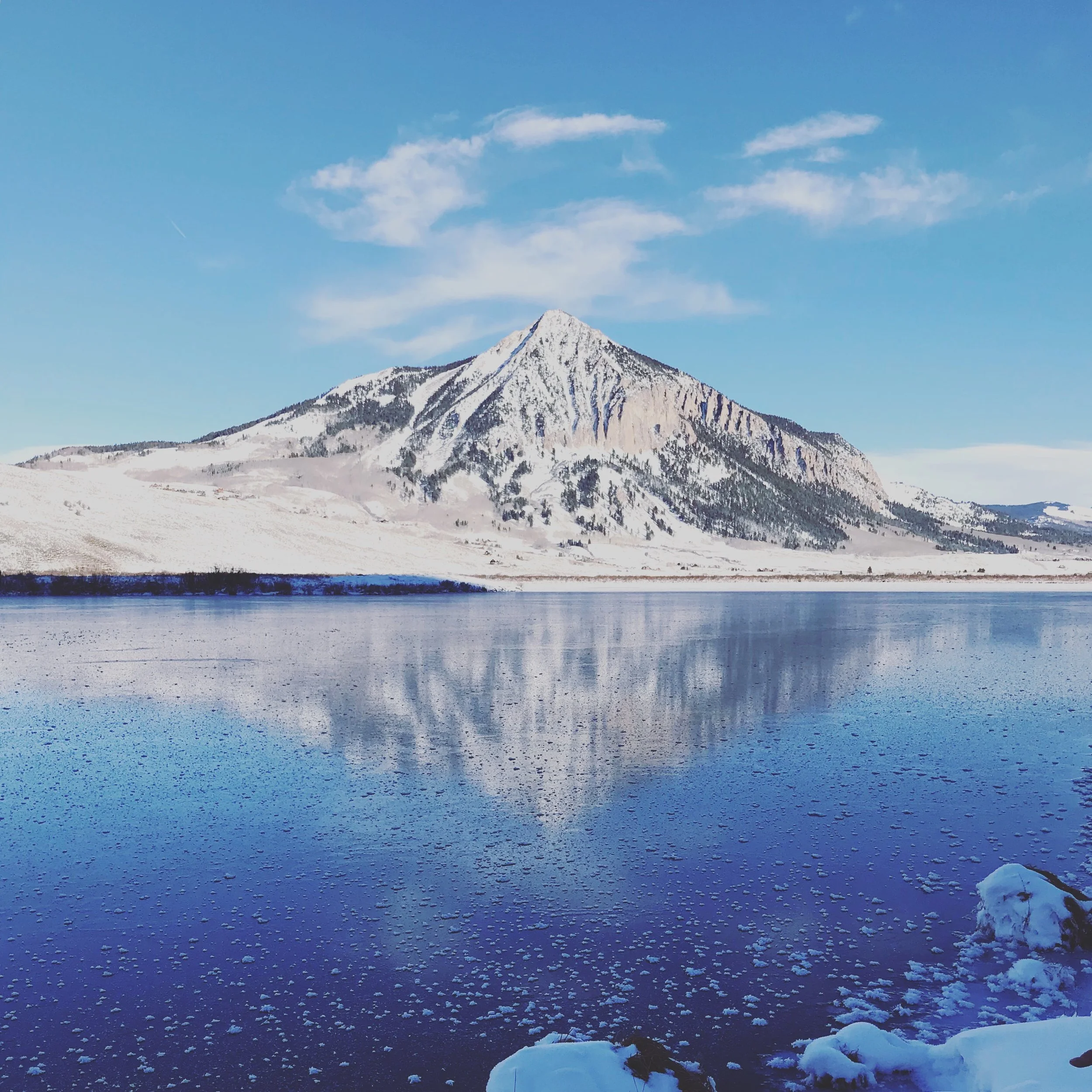 Snow-covered mountain with a partly frozen lake in the foreground under a blue sky with clouds.
