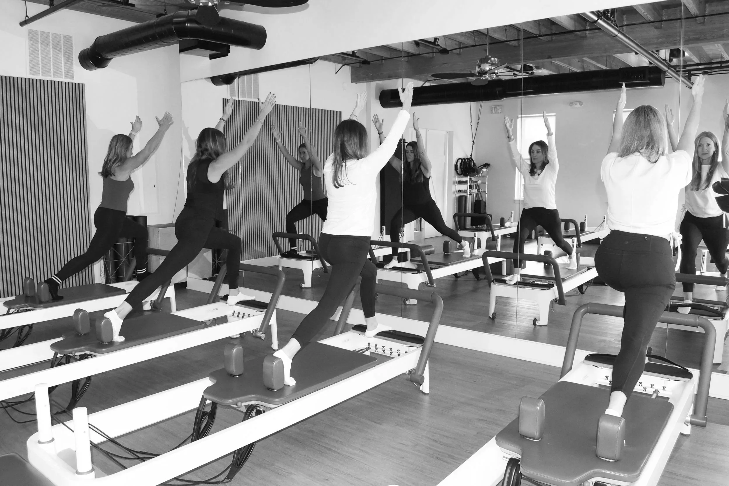 Group of women in athletic wear participating in a fitness class in a gym, performing stretching exercises on Pilates reformer machines, with a mirror reflecting their movements.