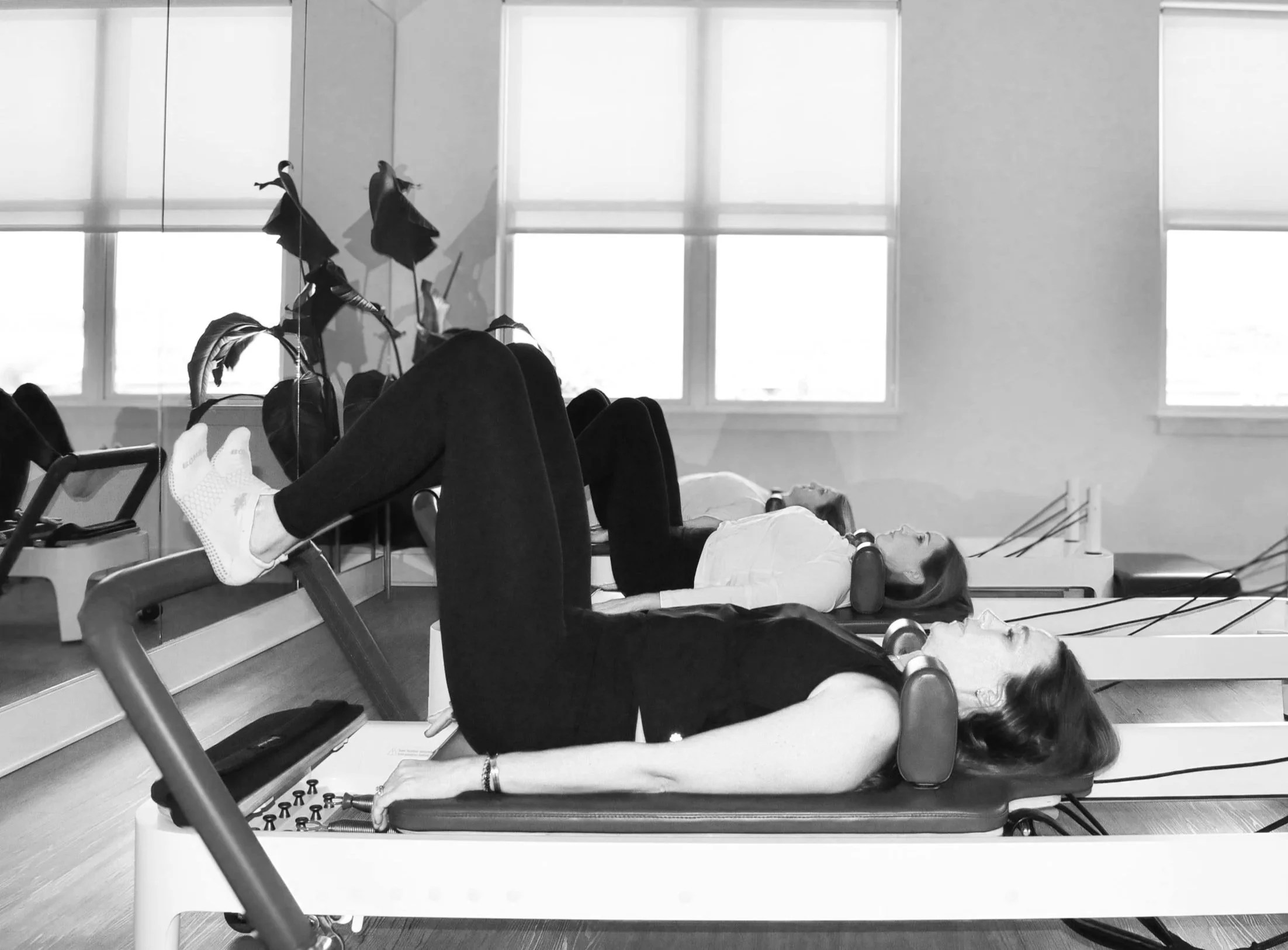 A person lying on their back on a medical examination table, with their arms at their sides and head turned to the side, in what appears to be a hospital or medical setting. There is another person partially visible at the foot of the table. The room has large windows with blinds and some medical equipment around.