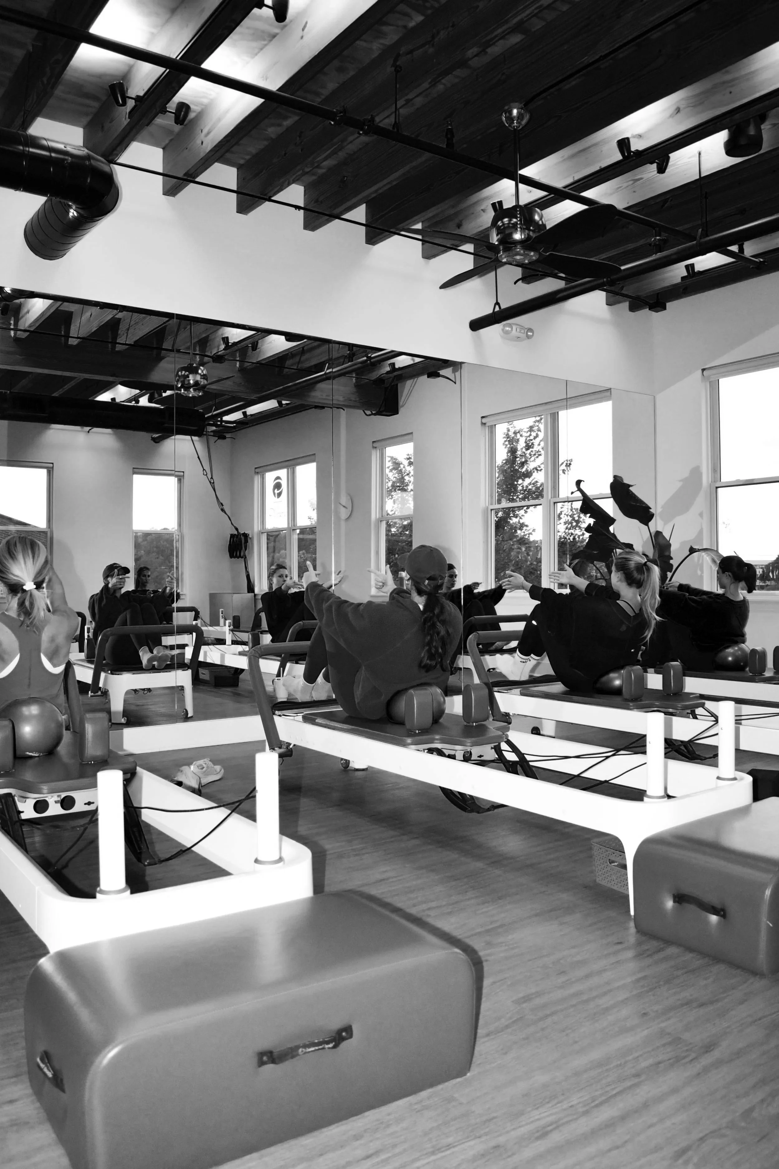 A group fitness class in a gym studio with women seated on Pilates reformer machines, extending their arms forward in a synchronized exercise. The studio has large windows, wooden ceiling beams, and an industrial-style ceiling with exposed pipes and ducts.