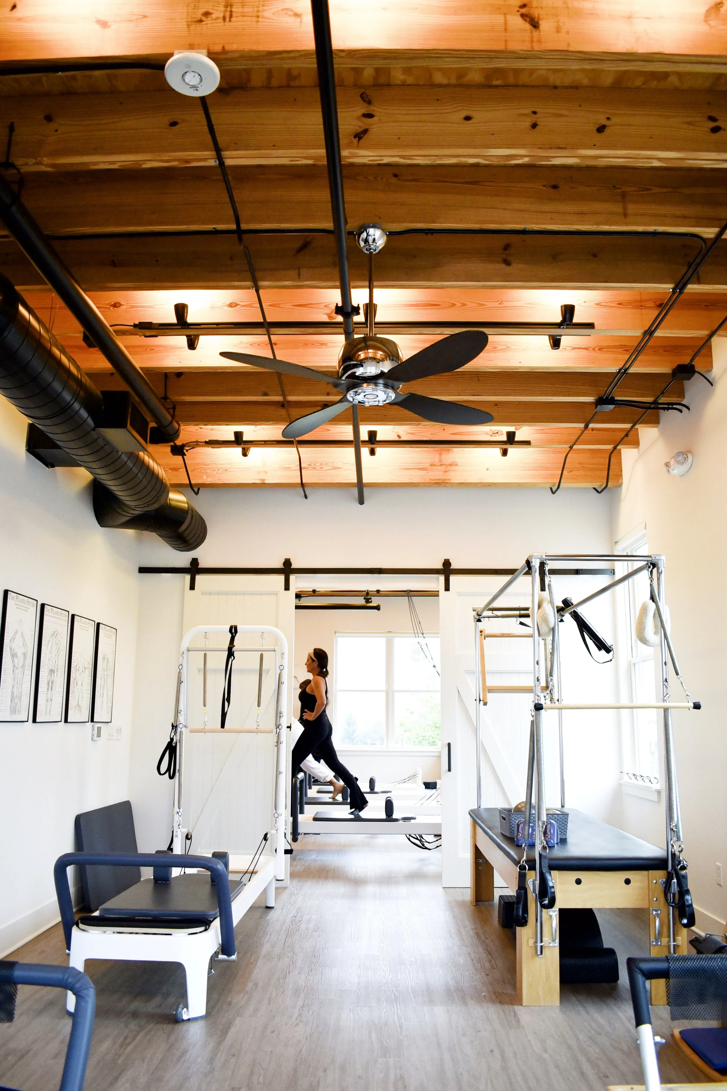 Interior view of a Pilates or fitness studio with wooden beams on the ceiling, black industrial pipes, and various exercise equipment including reformer machines, Pilates rings, and chairs; a woman standing on a reformer machine near the window.