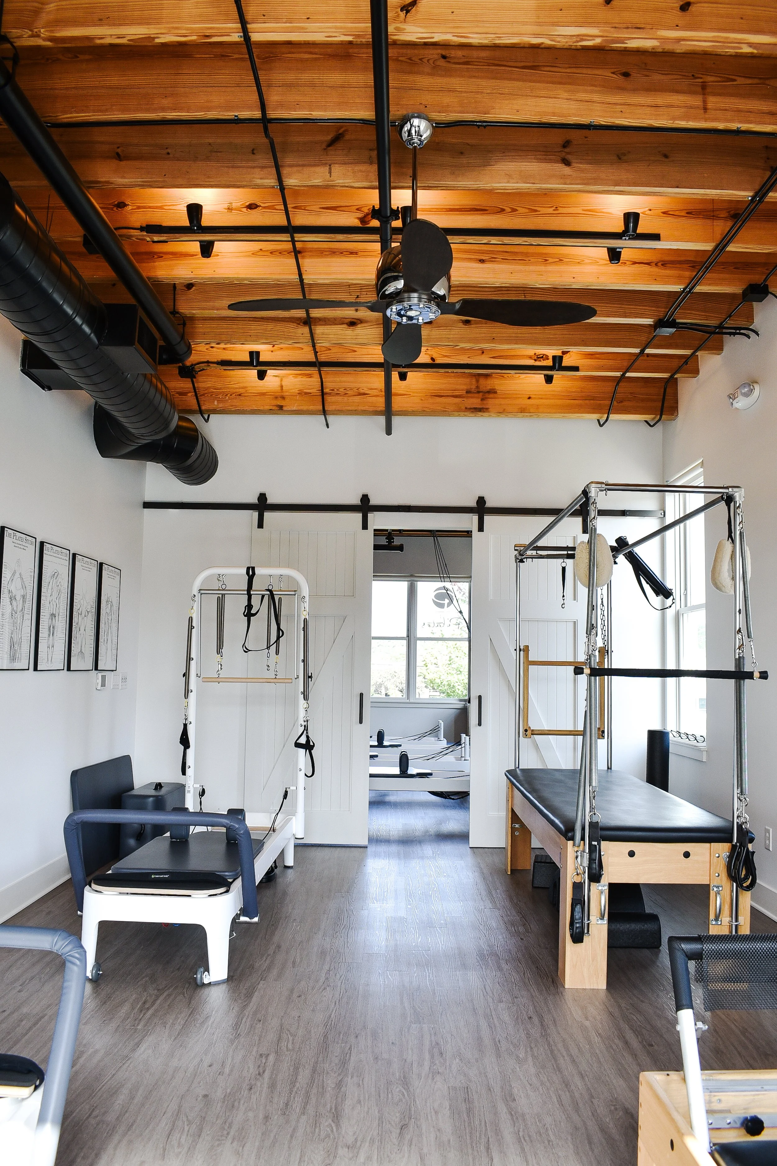 A Pilates or physical therapy studio with various exercise equipment, including a reformer, trapeze table, and other Pilates apparatus, with a wooden plank ceiling and large windows.