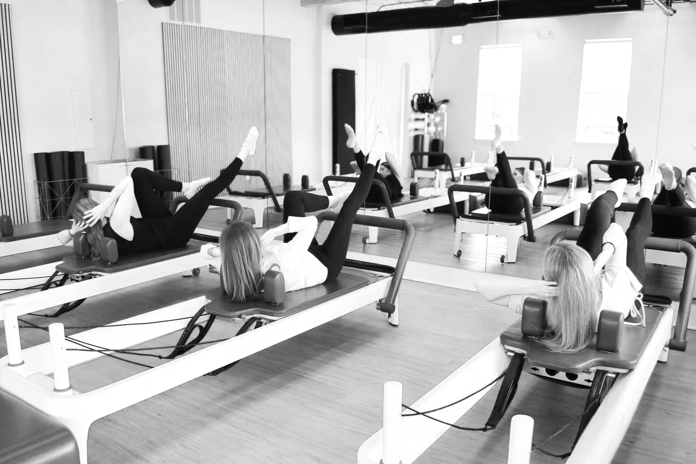 Women in a Pilates or yoga studio lying on reformer machines practicing leg stretches, with large windows in the background, black and white photo.