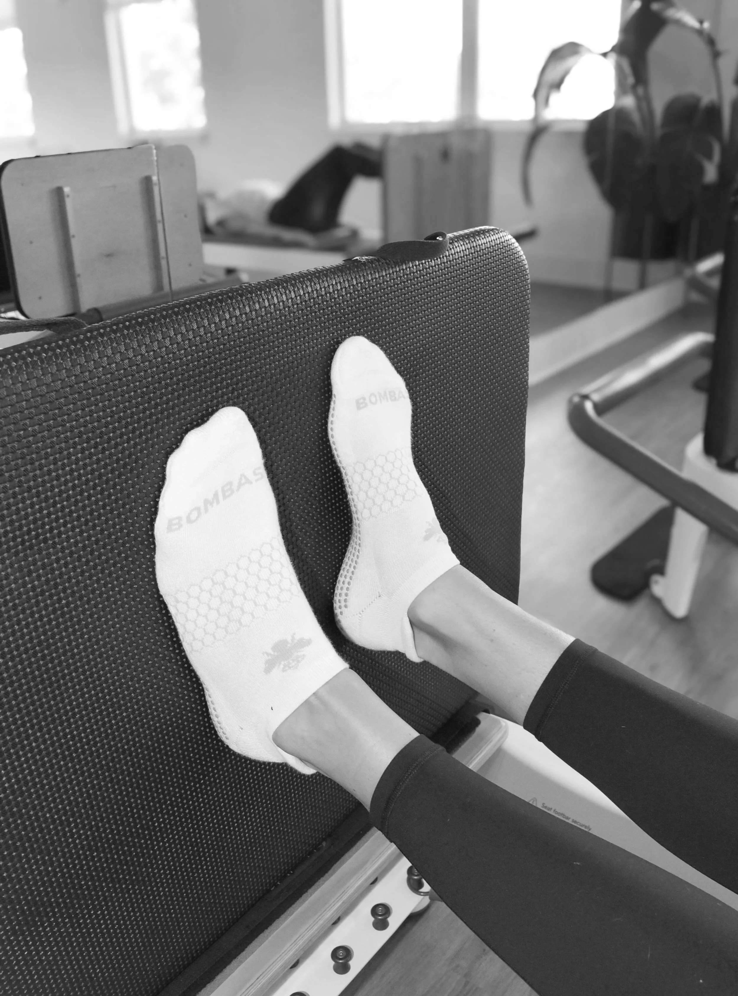Person wearing white sports socks with 'BOMBA' logo, lying on a piece of exercise equipment in a gym, with legs extended and hands doing leg raises.