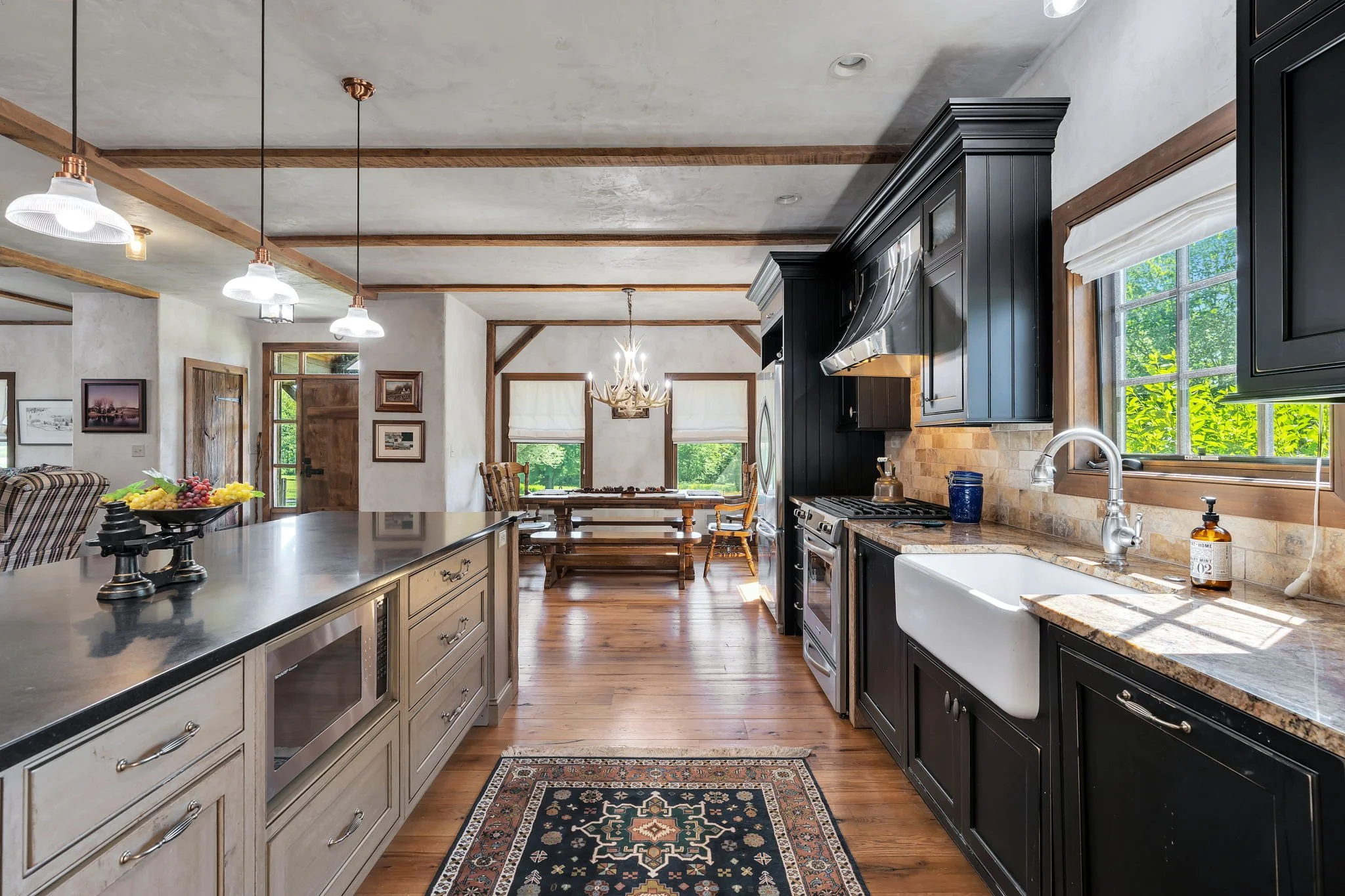 A spacious kitchen with a large island countertop, black cabinetry, wood accents, and modern pendant lighting. Visible dining area with wooden table and chairs, and a cozy living room with framed artwork on the walls.