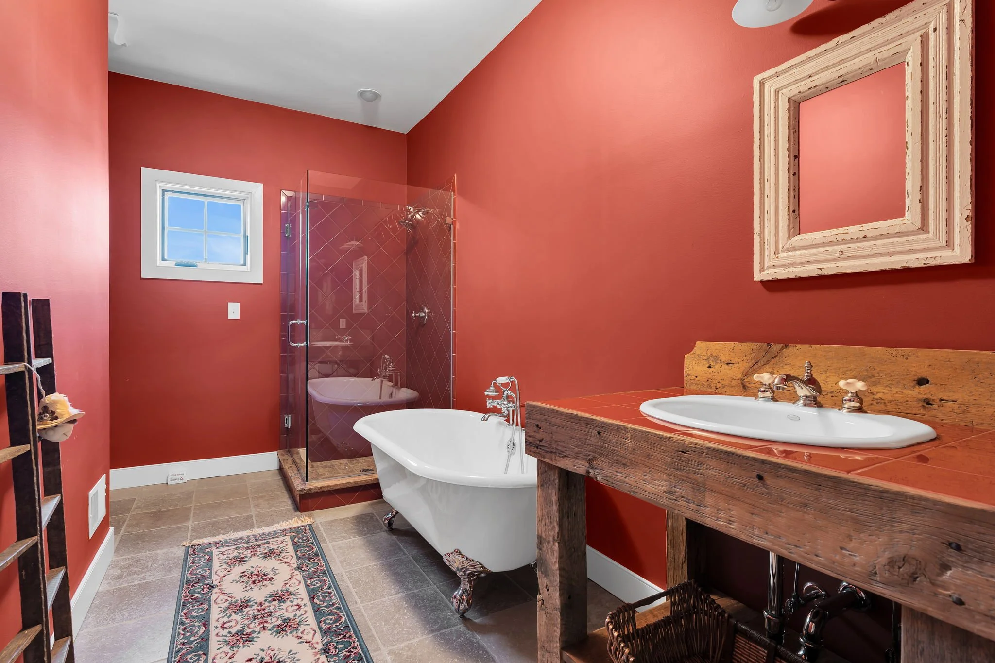 A rustic bathroom featuring a red wall and a clawfoot bathtub next to a wooden vanity with a white sink. There is a framed mirror on the wall, a small window, a tiled shower area, and decorative rugs on the tile floor.