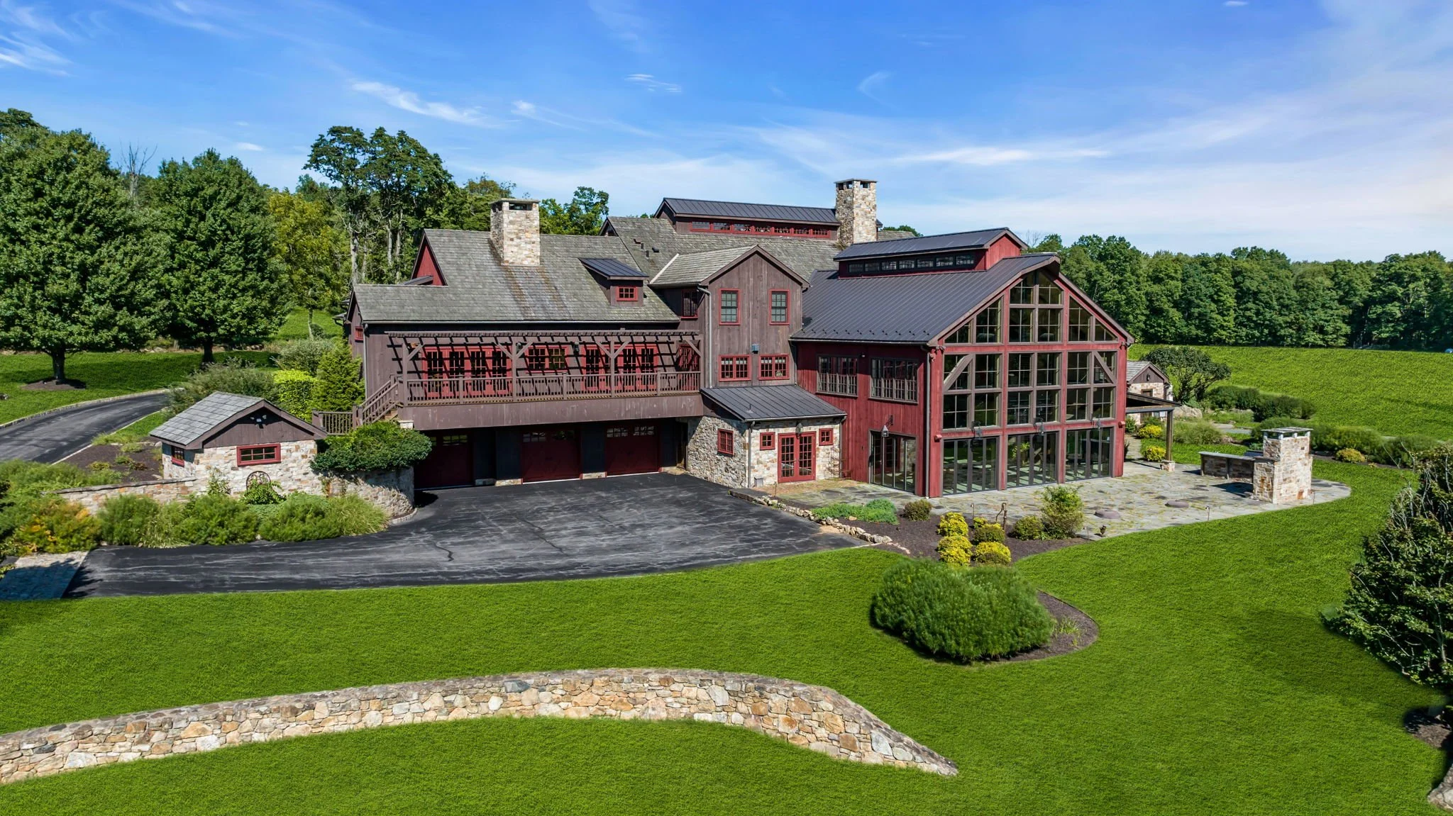 A large, rustic house with red wooden siding and multiple stone chimneys, surrounded by landscaped green lawns and trees, with a bright blue sky overhead.