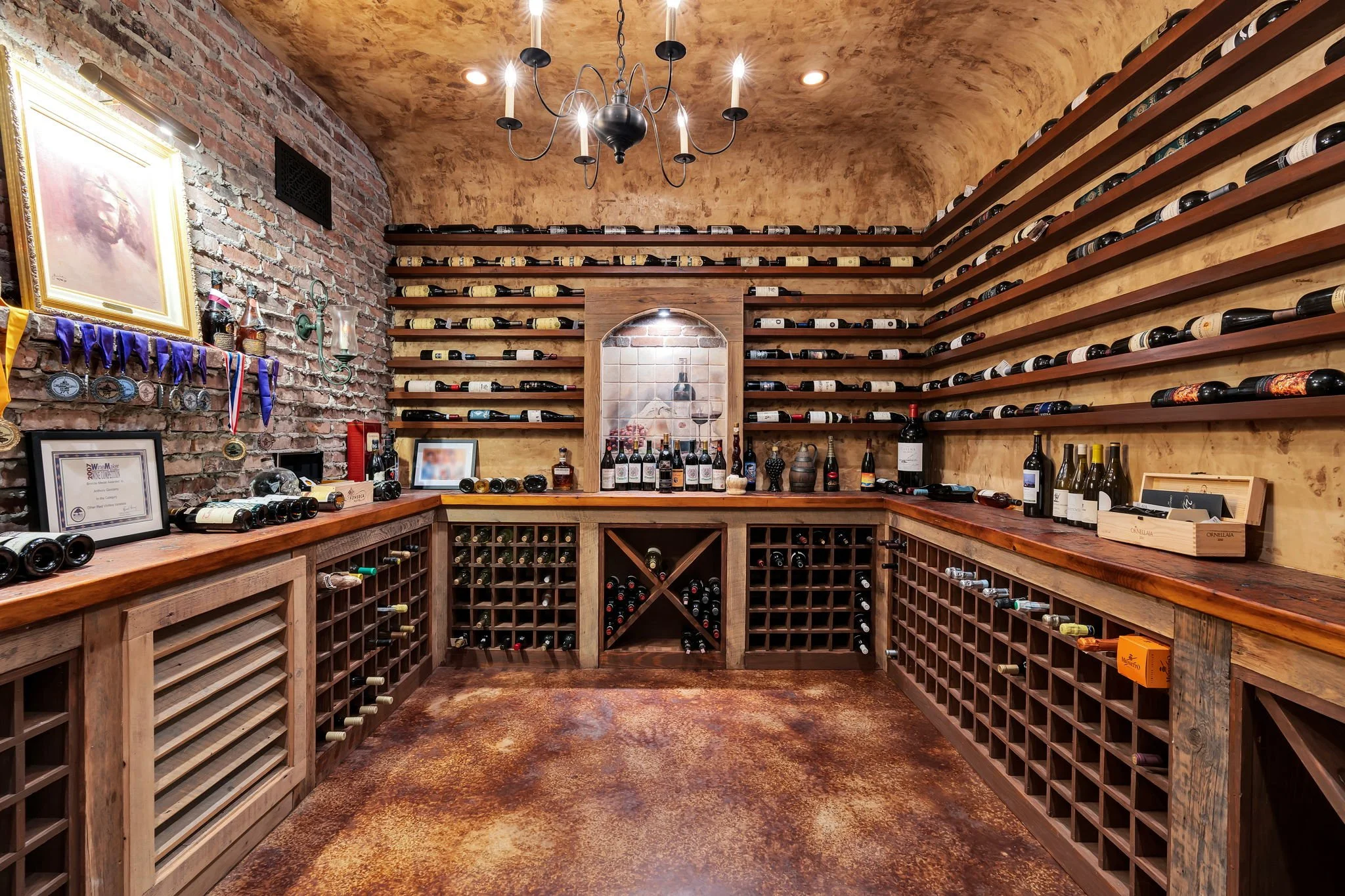 A wine cellar with wooden wine racks filled with wine bottles, exposed brick wall, a chandelier hanging from the ceiling, and a countertop with more wine bottles, awards, and framed pictures.