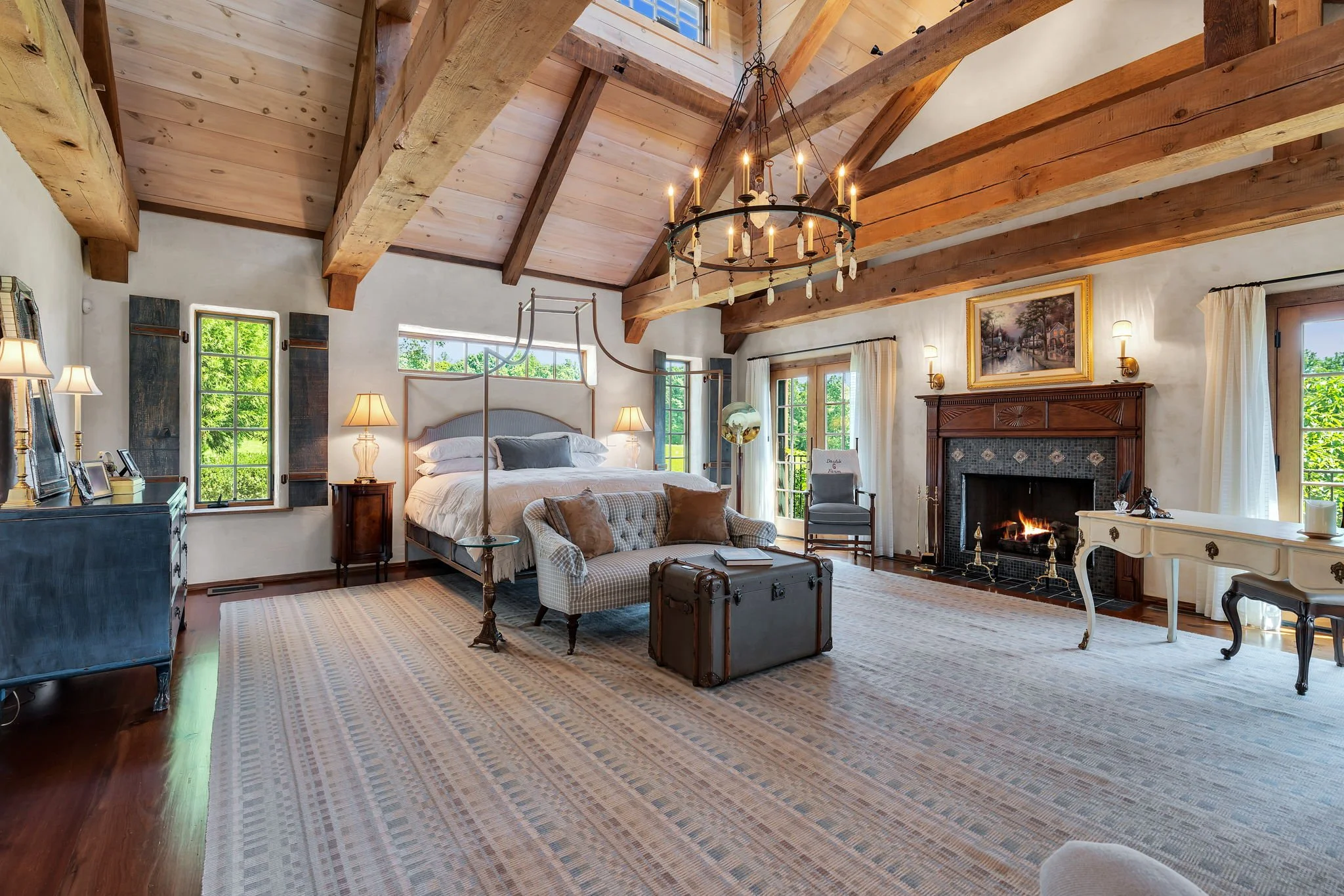 A spacious living room with a high, vaulted wooden ceiling, exposed wooden beams, and large windows revealing greenery outside. The room features a fireplace with a wooden mantel, a chandelier hanging from the ceiling, a bed with white linens and pil