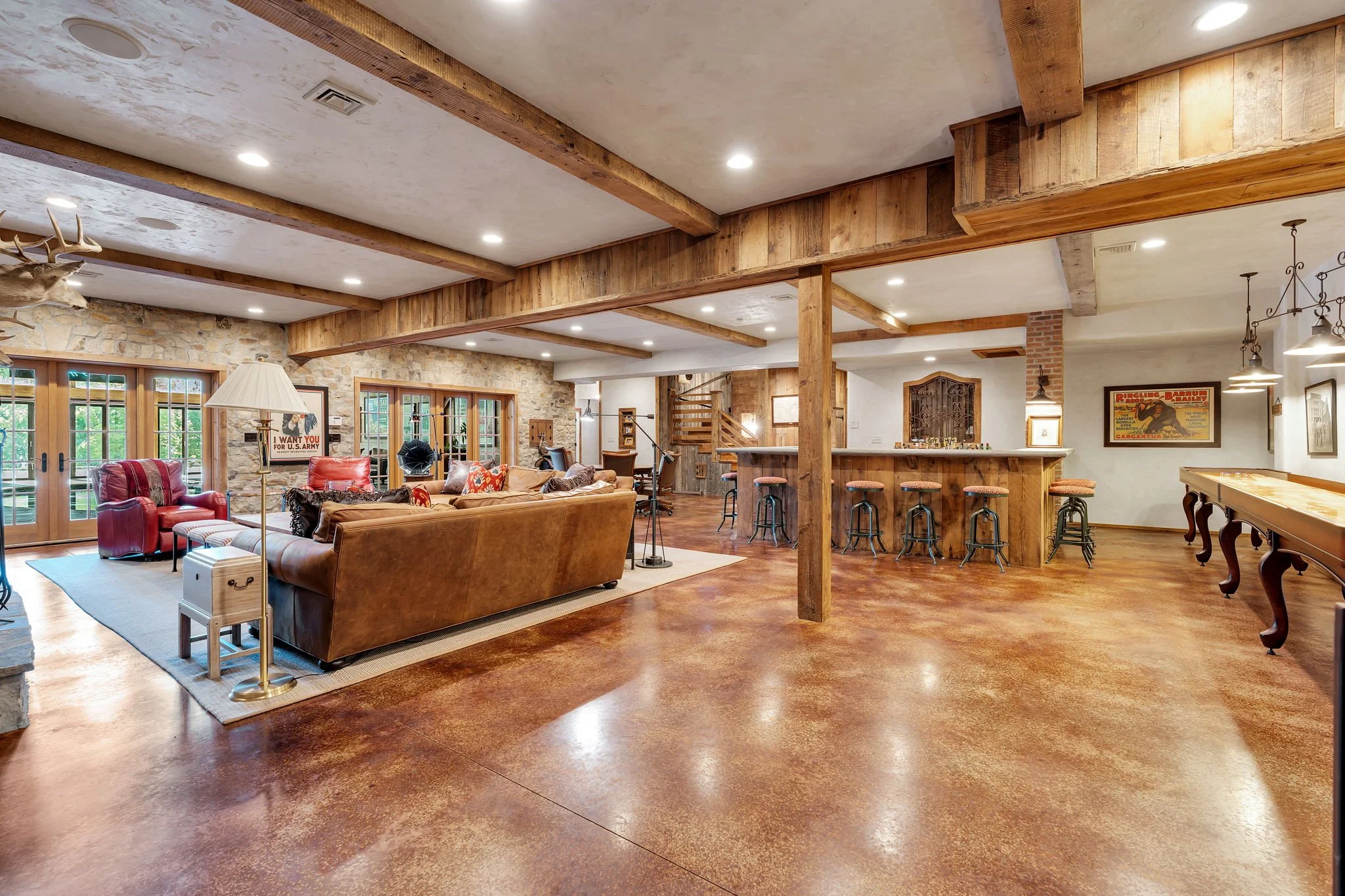 A spacious living area with a rustic design featuring wooden beams on the ceiling, stone walls, large windows with wooden frames, a brown leather sofa, red chairs, a side table, and a patterned area rug. There is a bar area with stools, framed poster
