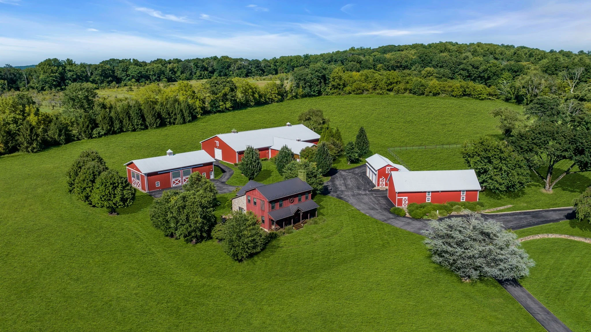 Aerial view of a rural farm with red barns, surrounded by green fields and trees, and a wooded hillside in the background under a blue sky.