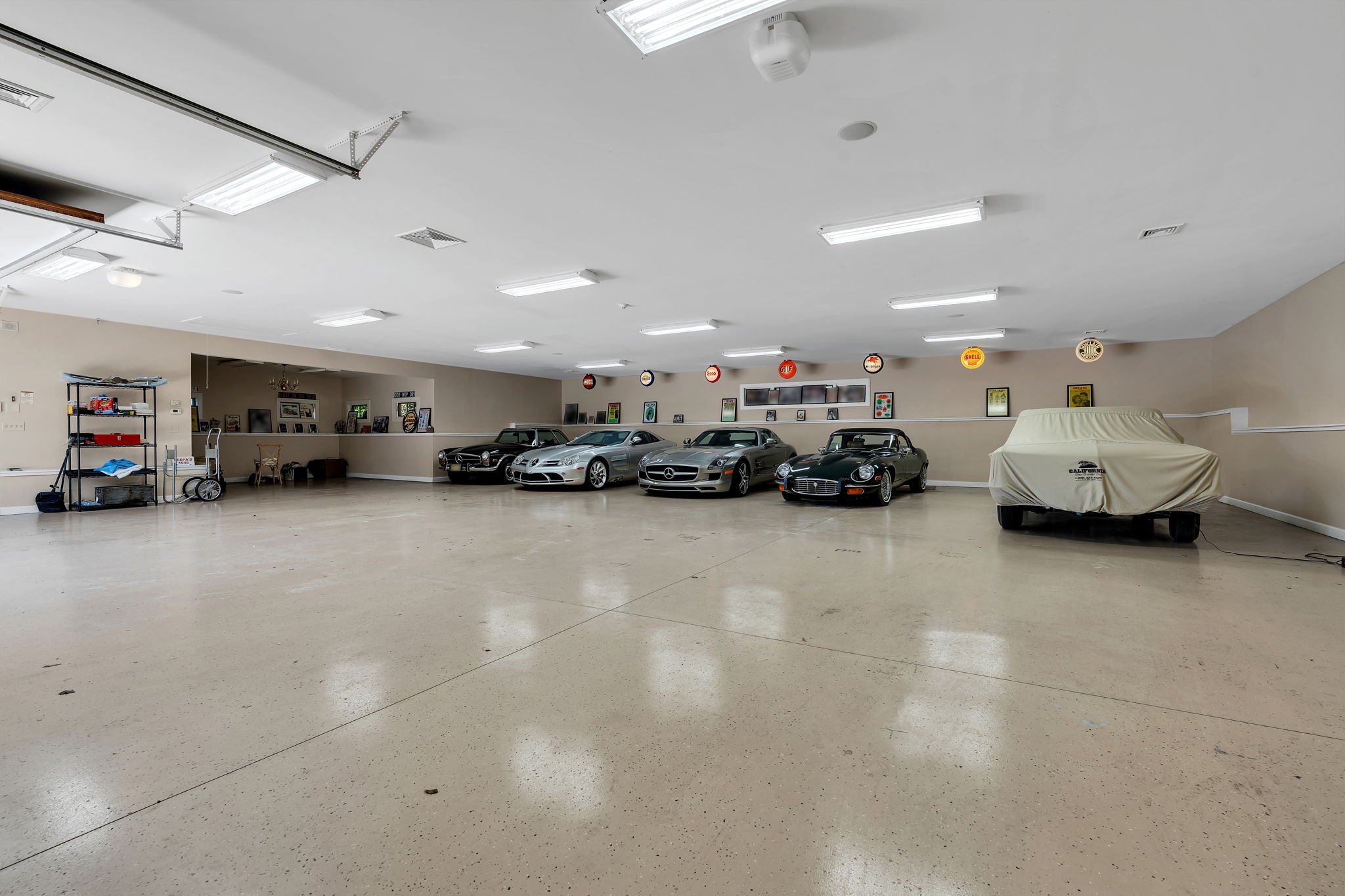 Empty garage with four luxury cars parked along a beige wall, vintage signs hanging above, and a covered vehicle with a California license plate.