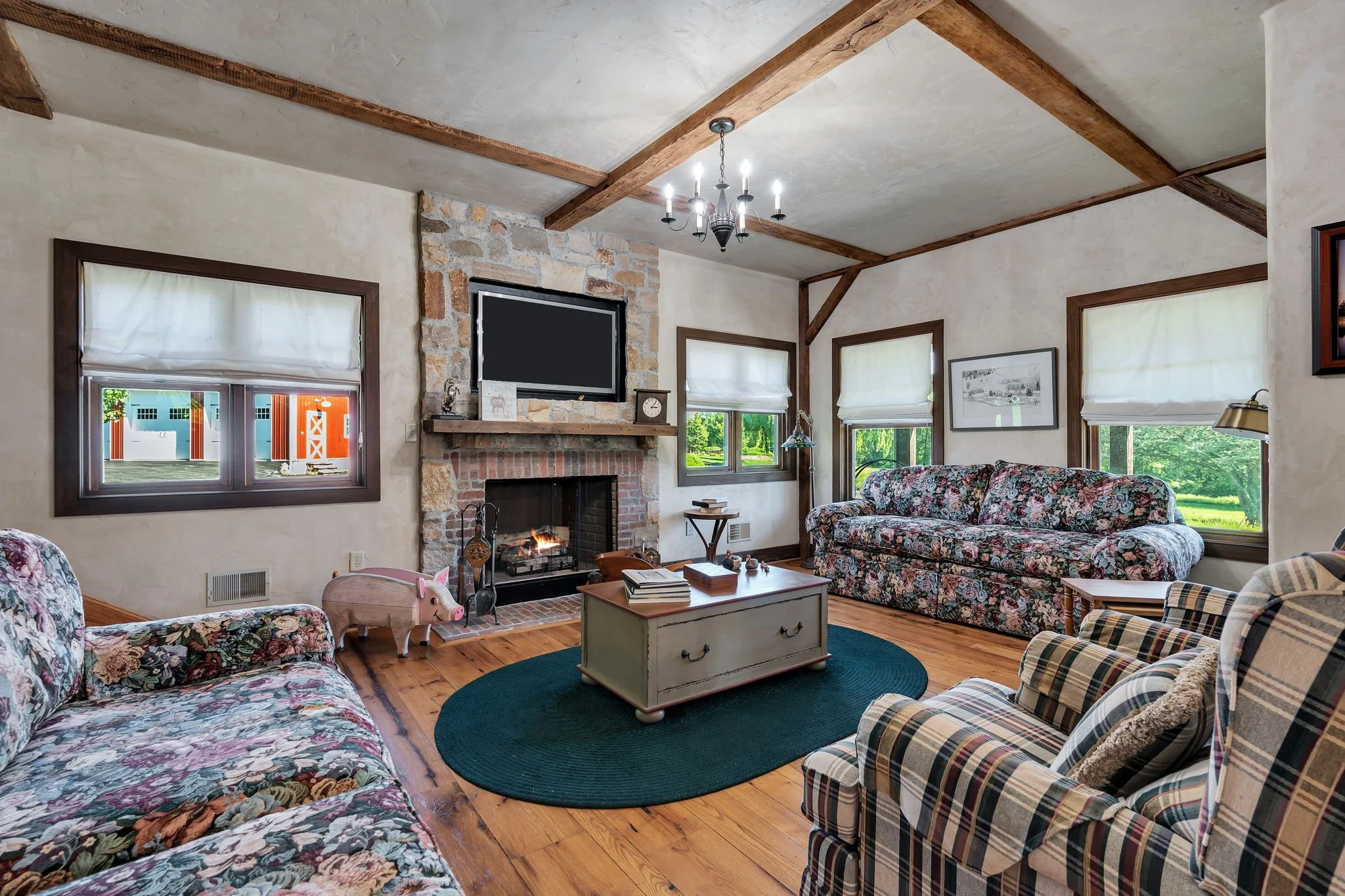 Living room with floral sofas, plaid armchair, coffee table, fireplace with TV above, stone chimney, windows with white blinds, hardwood floors, and wooden beams on the ceiling. There is a chandelier hanging from the ceiling.