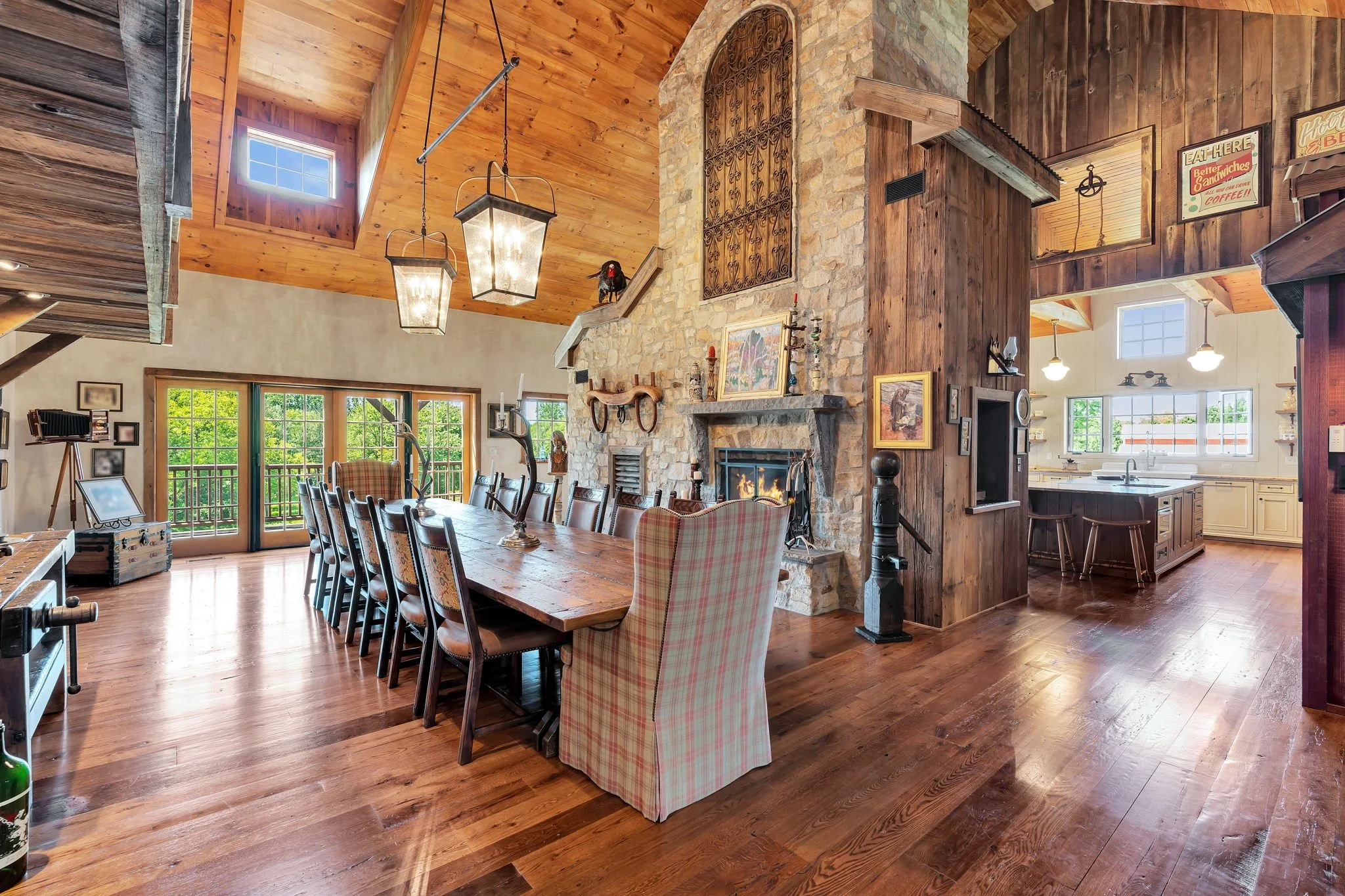 Open concept living and dining room with wood-paneled vaulted ceiling, large windows with greenery outside, a stone fireplace, and a wooden dining table with plaid armchairs.