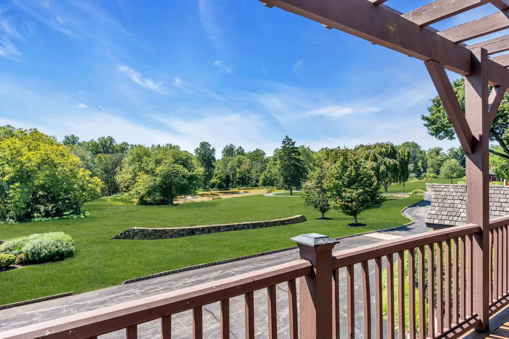 View from a wooden balcony overlooking a well-maintained grassy area with trees, a winding pathway, and a stone bench under a clear blue sky with some clouds.