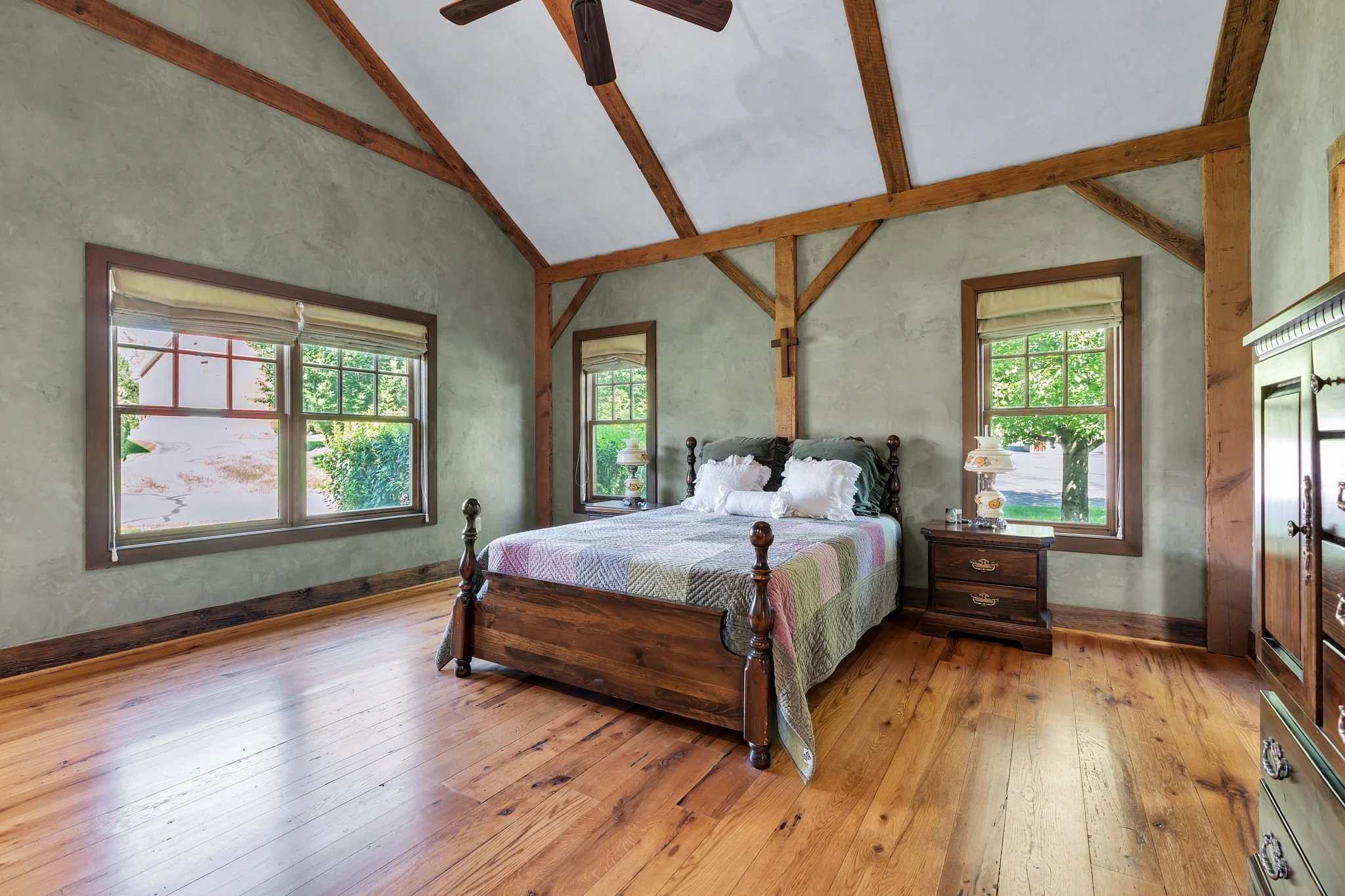 A cozy bedroom with wooden beams on the ceiling, three windows with green foliage outside, a wooden bed with white bedding, a nightstand with a lamp, and a wooden dresser.