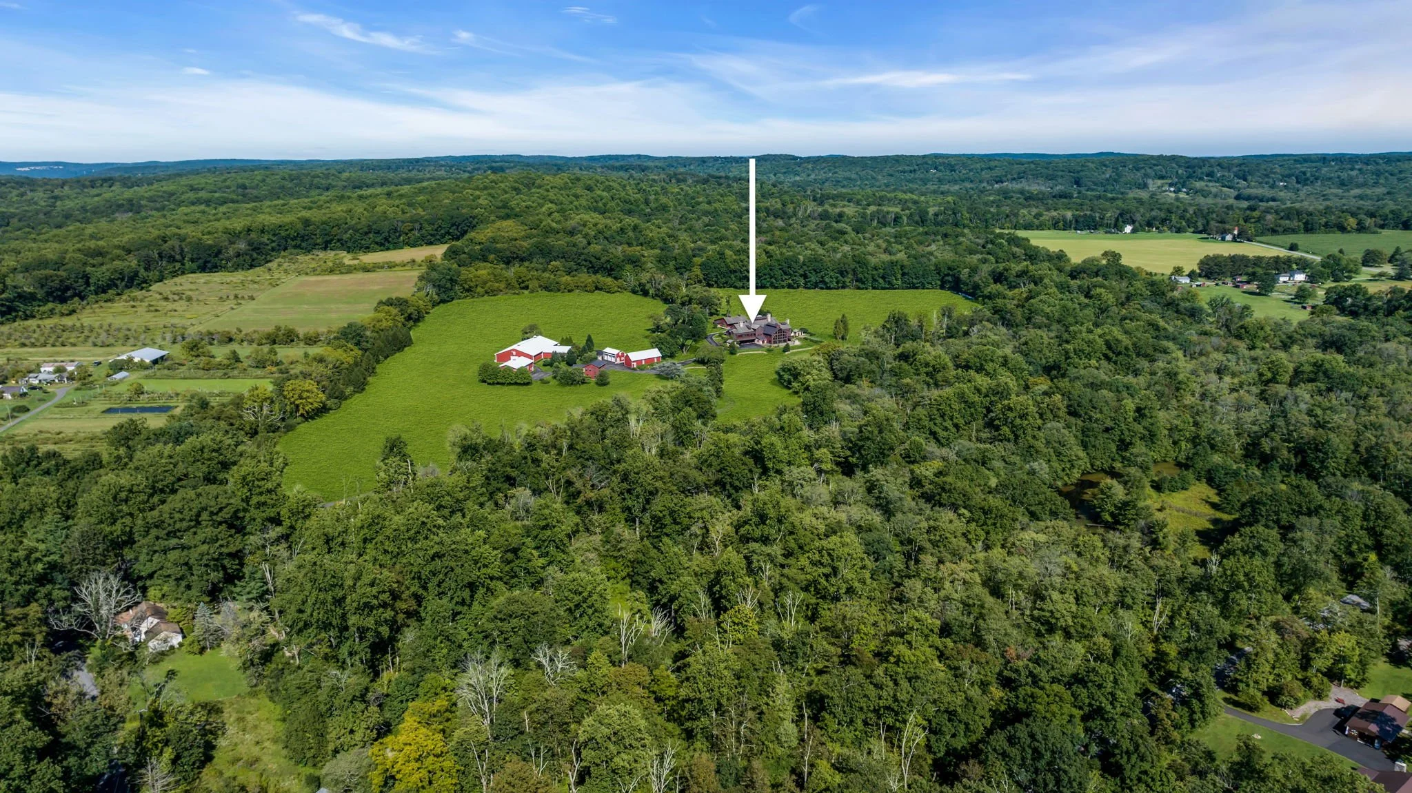Aerial view of a large house with a dark roof surrounded by trees and green fields, with rolling hills and a blue sky in the background.