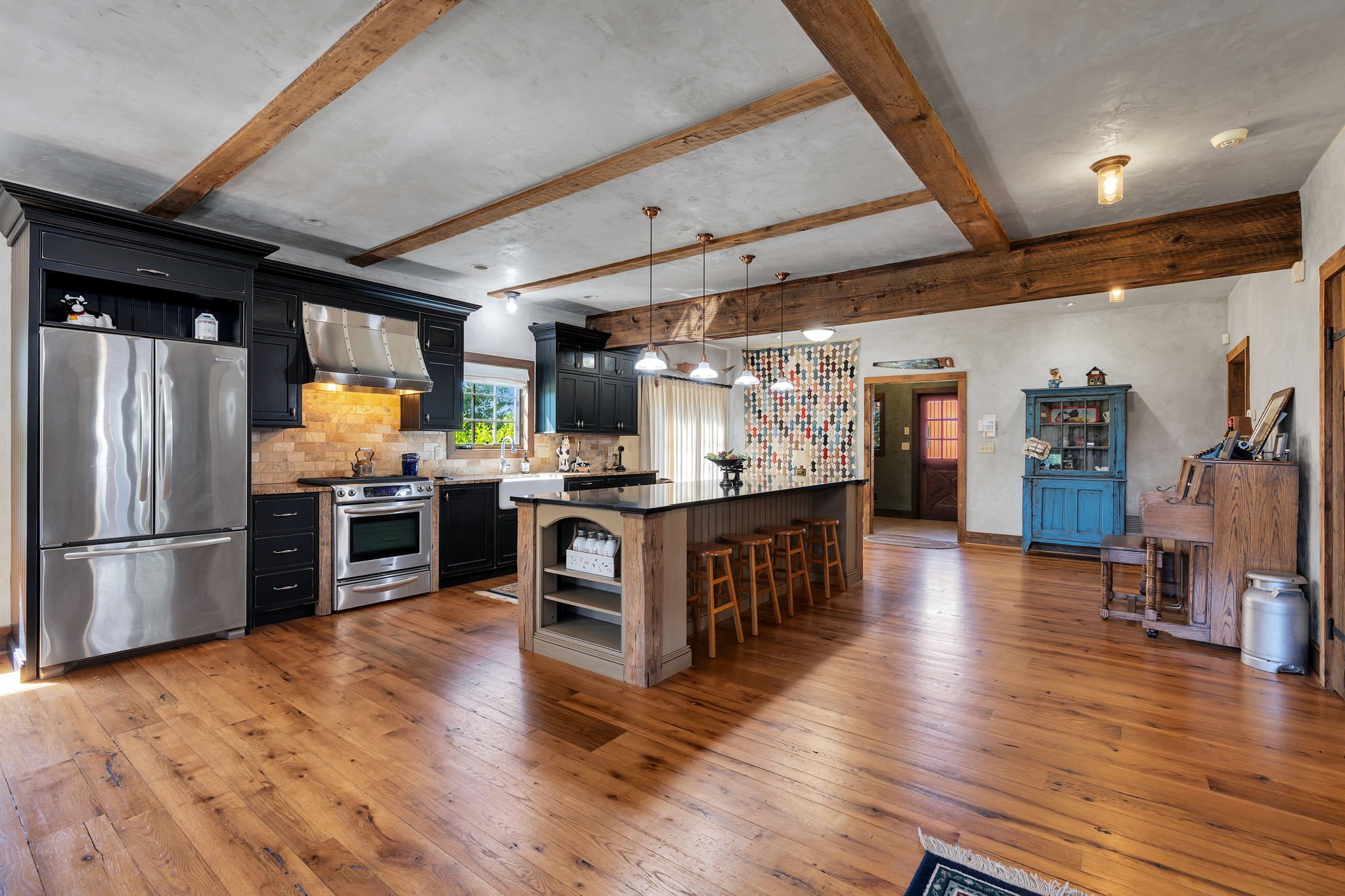 Open-concept kitchen with wooden flooring, black cabinets, stainless steel refrigerator, oven, and range hood. A kitchen island with bar stools, cozy seating area with a blue cabinet, and warm lighting.