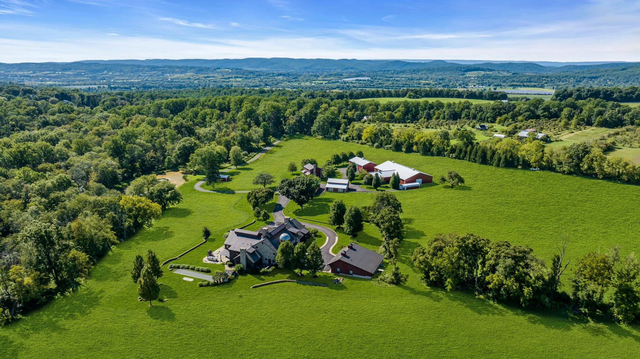 An aerial view of a rural landscape with lush green fields, trees, and a few houses and barns, under a blue sky with scattered clouds.