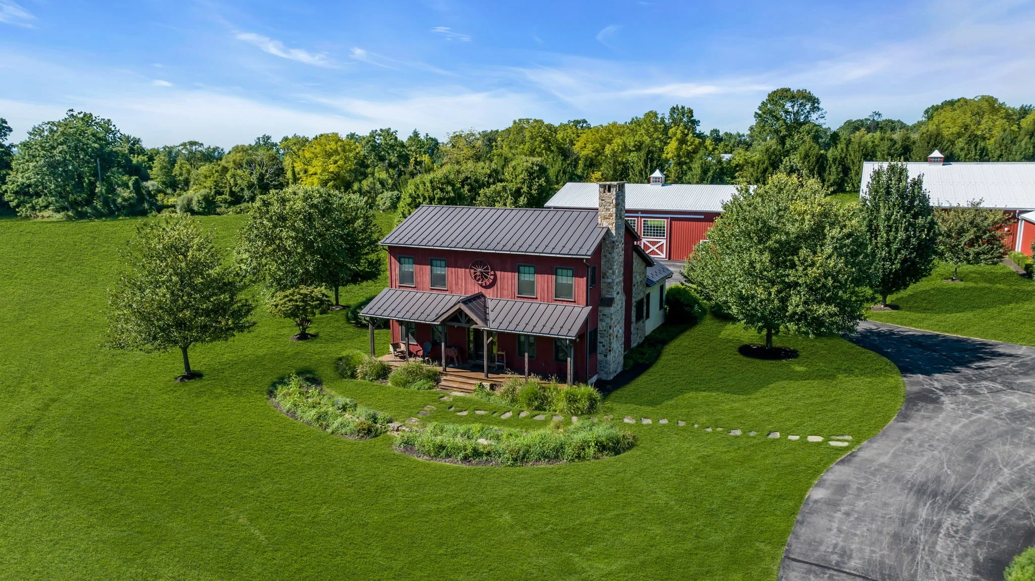 A two-story red house with a metal roof, a stone chimney, and front porch, surrounded by green grass and trees, with a driveway on the right and a background of wooded area under a clear blue sky.