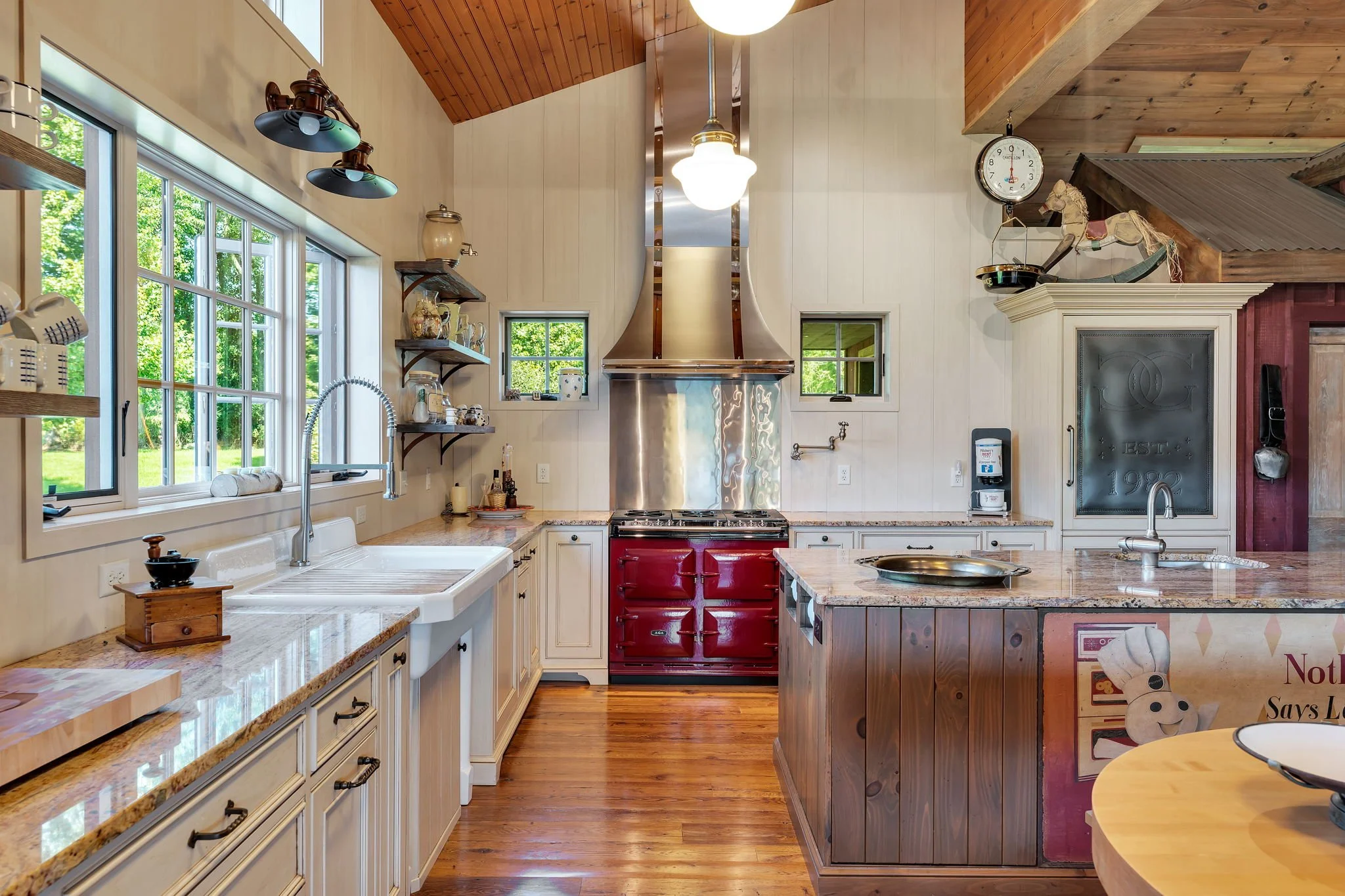 Kitchen with white cabinets, granite countertops, stainless steel range hood, red oven, and wooden accents. Large windows with greenery outside, decorative items on shelves, ceiling lights, and hardwood floors.