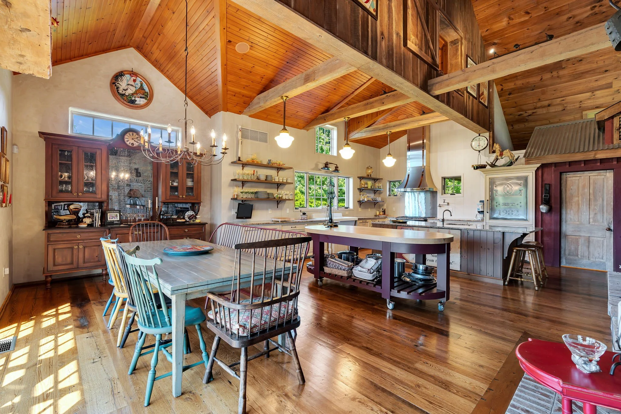 The interior of a rustic kitchen and dining area with wood-paneled vaulted ceilings, a chandelier, open shelves, a kitchen island, and mixed vintage and modern furniture and decor.