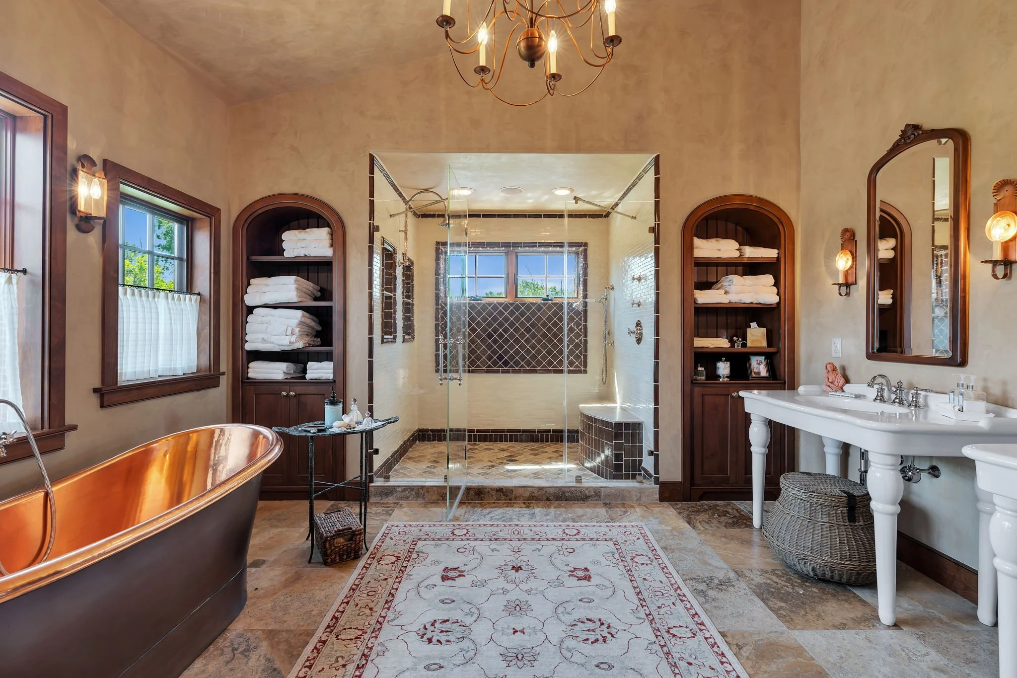A spacious bathroom featuring a copper bathtub, a glass-enclosed shower with brown tiling, wooden cabinets filled with white towels, a white double sink vanity with a large mirror, and a decorative rug on the tiled floor. The room is well-lit with na