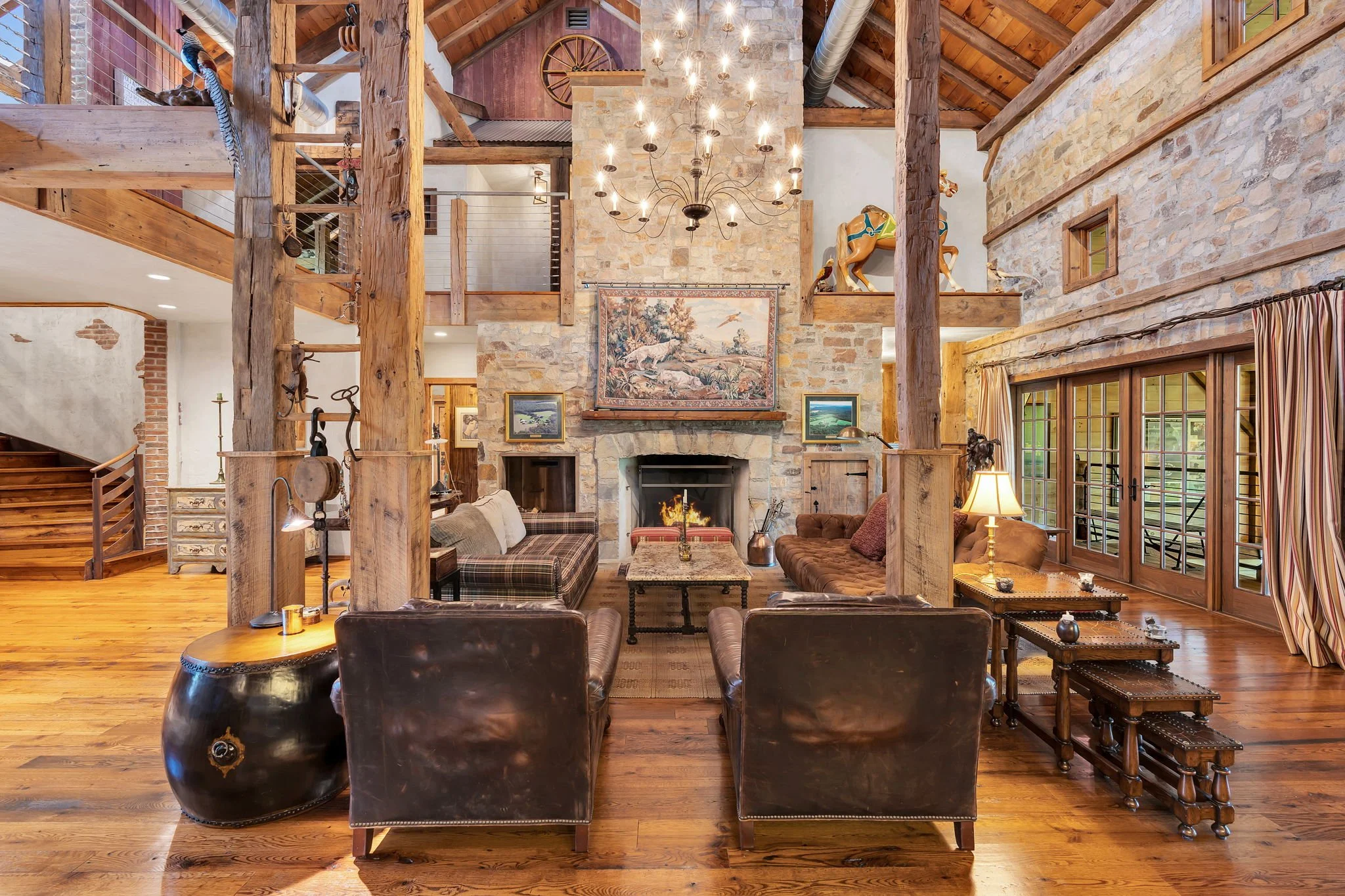 A spacious living room with a stone fireplace, wooden beams, and a high ceiling. There are leather chairs and a sofa around a coffee table. A chandelier hangs from the ceiling, and artwork decorates the walls. Large windows with striped curtains let 