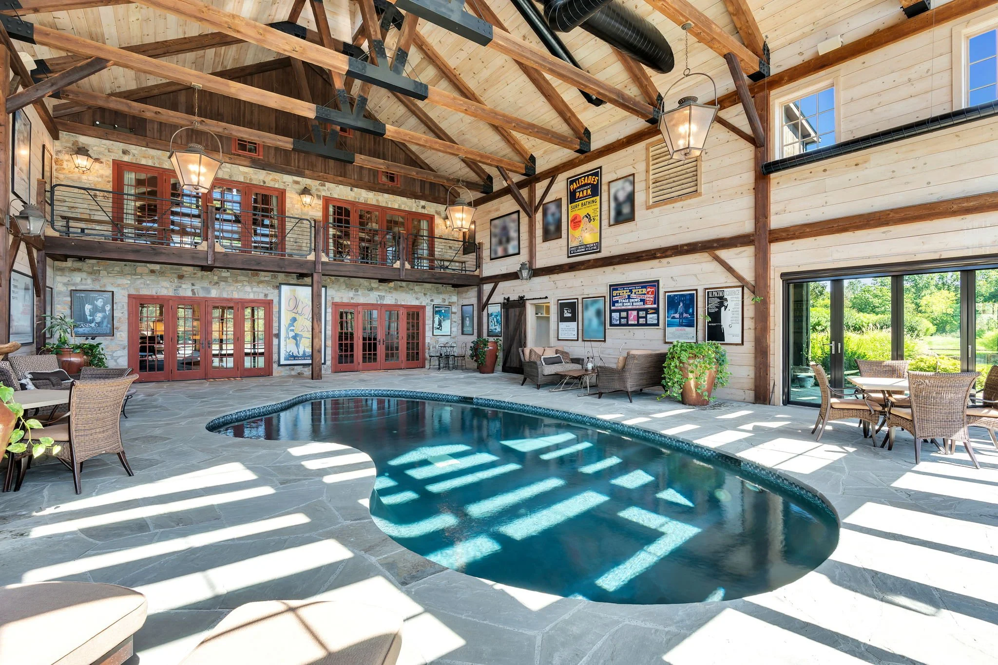 Indoor pool area with natural wood vaulted ceiling, exposed beams, and large windows. The space has seating with wicker chairs, framed artwork on the walls, and a patio door leading outside. There's also a balcony overlooking the pool and a brick wal