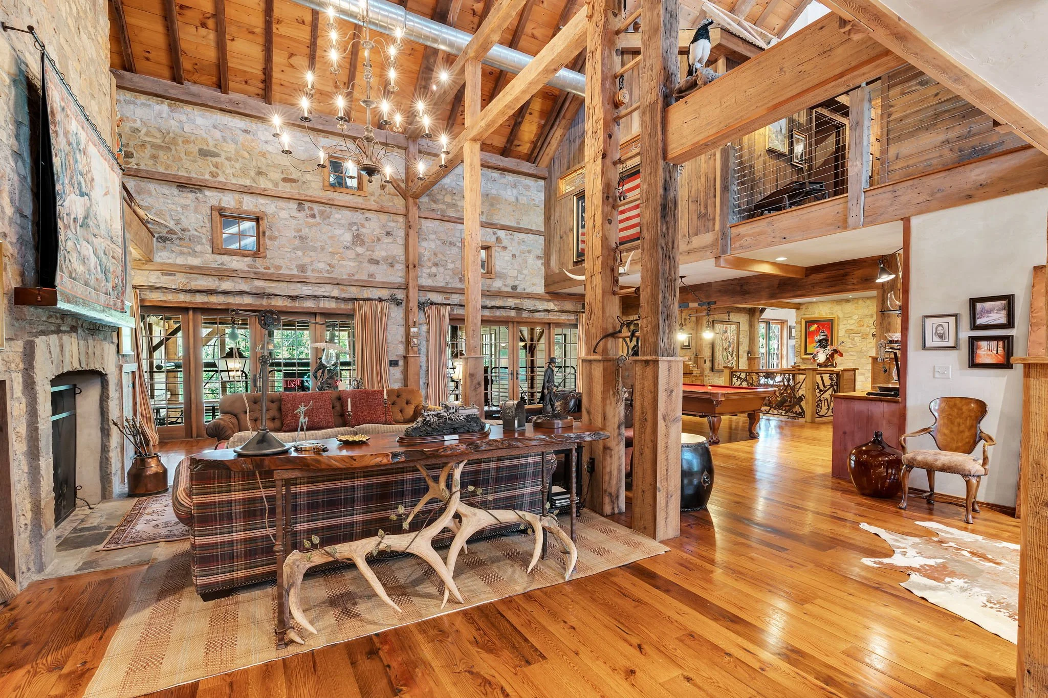 The image shows the interior of a rustic-style home with exposed wooden beams, stone walls, and wooden flooring. There is a large chandelier hanging from the ceiling and various furniture pieces including a plaid sofa, wooden coffee table, and decora