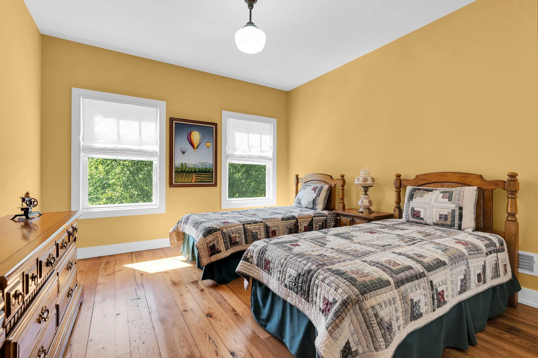 A bedroom with two twin beds with patchwork quilts, a wooden dresser, two windows with white blinds, yellow walls, a framed painting of hot air balloons, and a ceiling light.