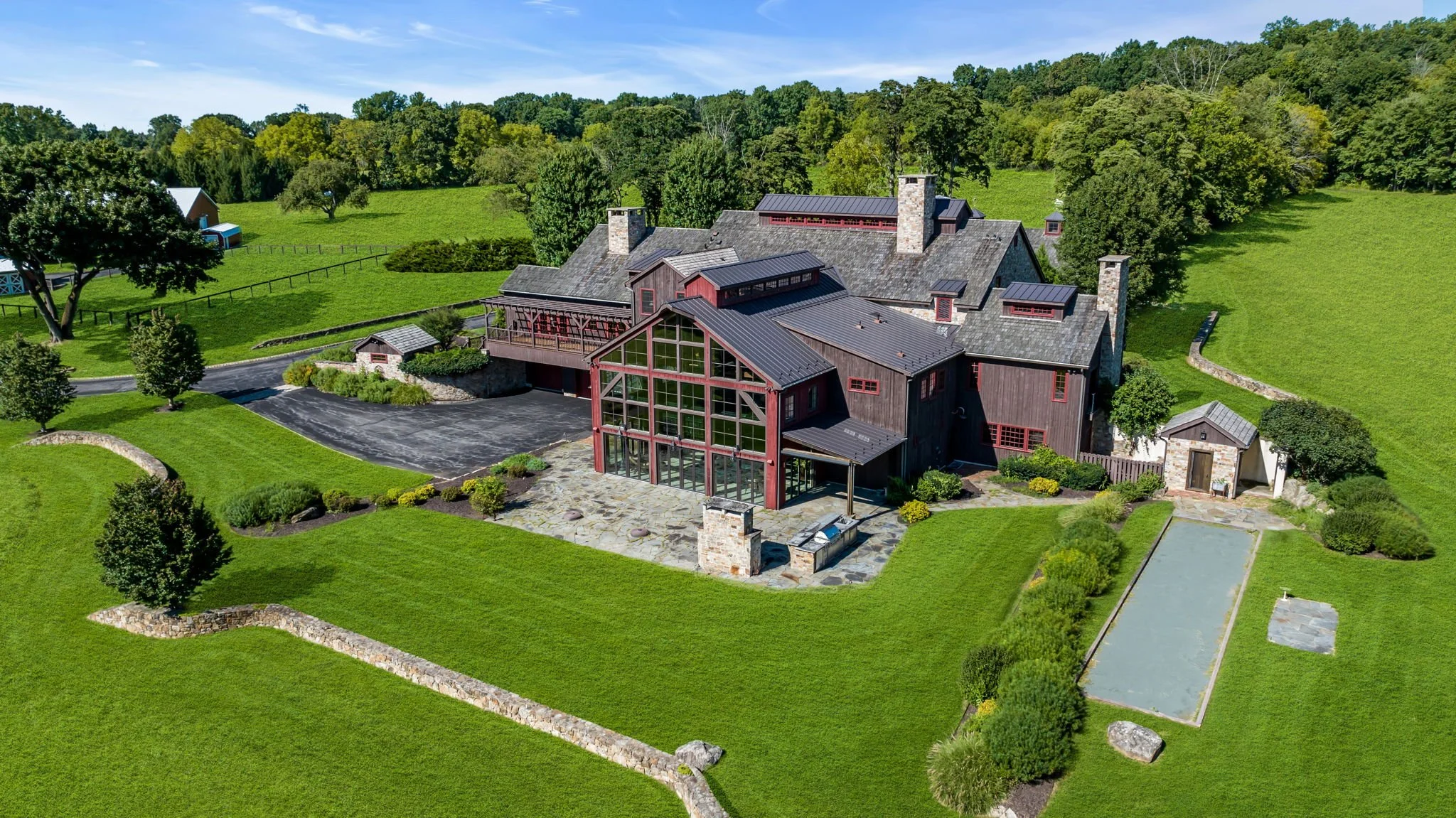 A large, modern house with a combination of stone and wood exterior, multiple chimneys, and a metal roof, surrounded by extensive green lawns, trees, and landscaping, with a tennis court and a driveway.