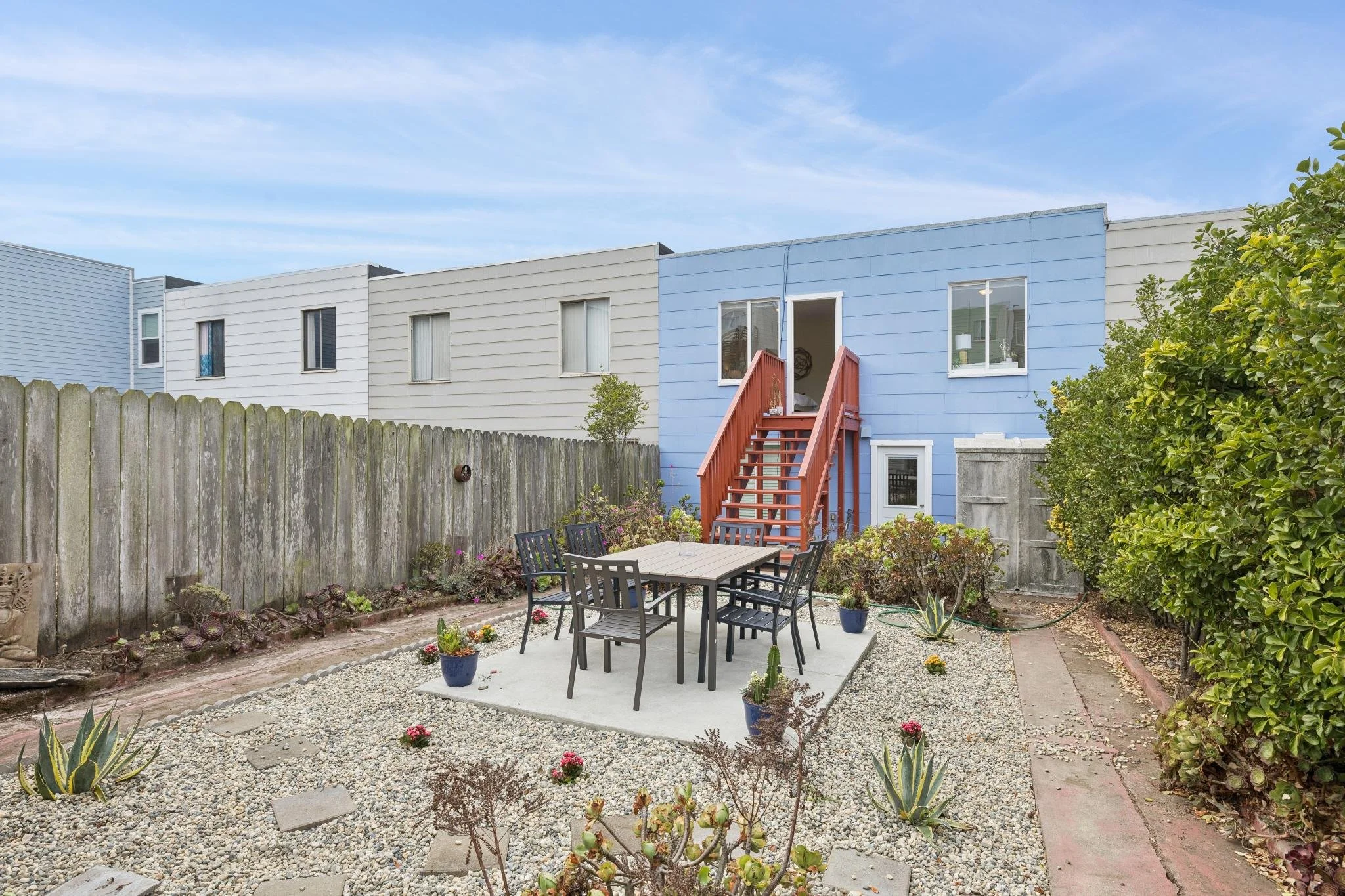 A backyard patio with a black outdoor dining table and chairs, surrounding plants, a gravel ground, a wooden fence, and colorful houses in the background with blue and beige siding, set against a partly cloudy sky.
