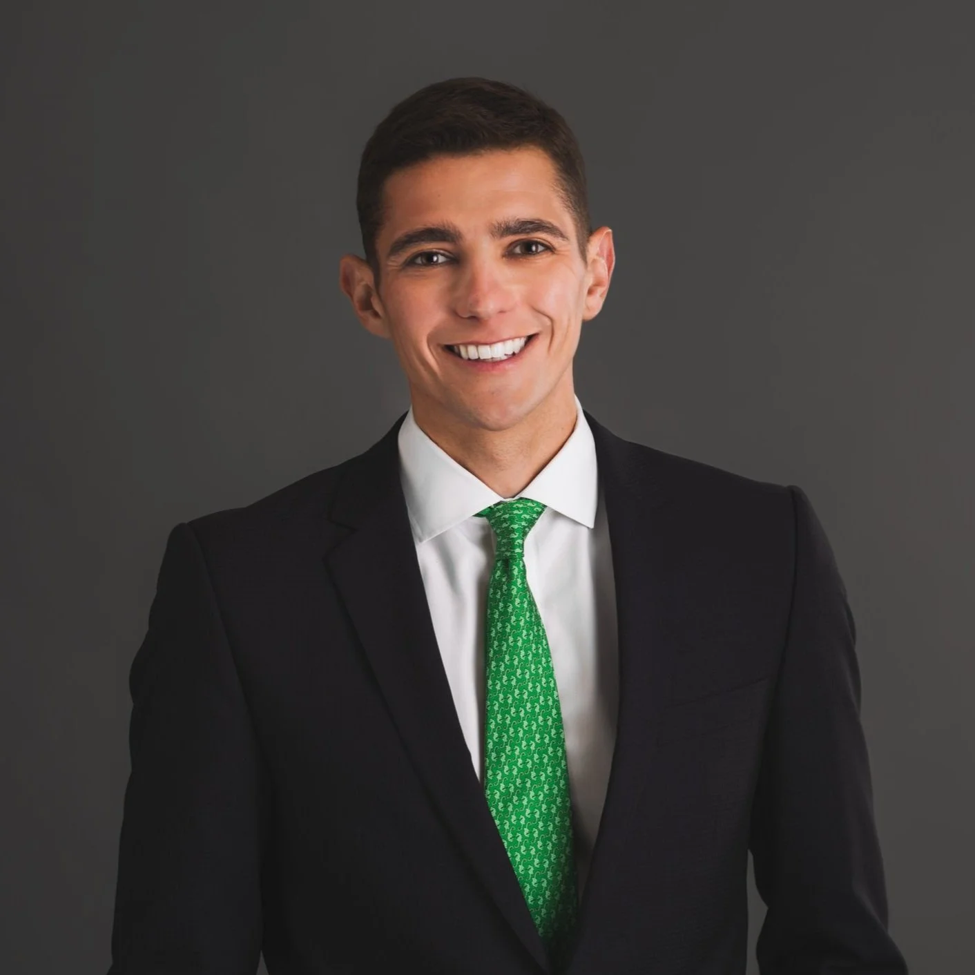 A young man with short dark hair smiling, wearing a black suit, white shirt, and green tie, against a gray background.