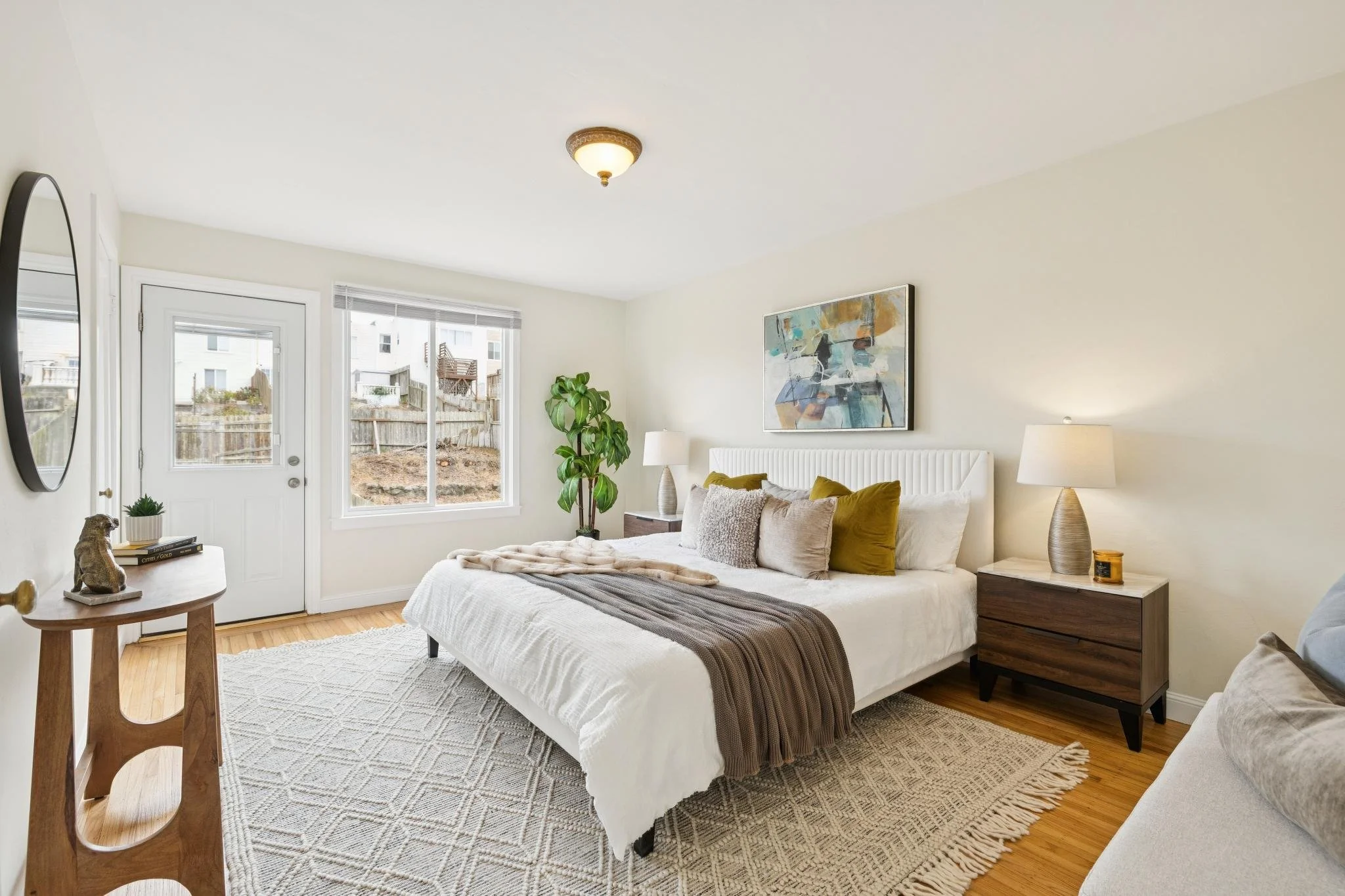 Bright bedroom with white walls, a large bed with beige and mustard pillows, a wooden side table with a lamp, a framed abstract painting on the wall, a potted plant near the window, and a sliding glass door leading outside.