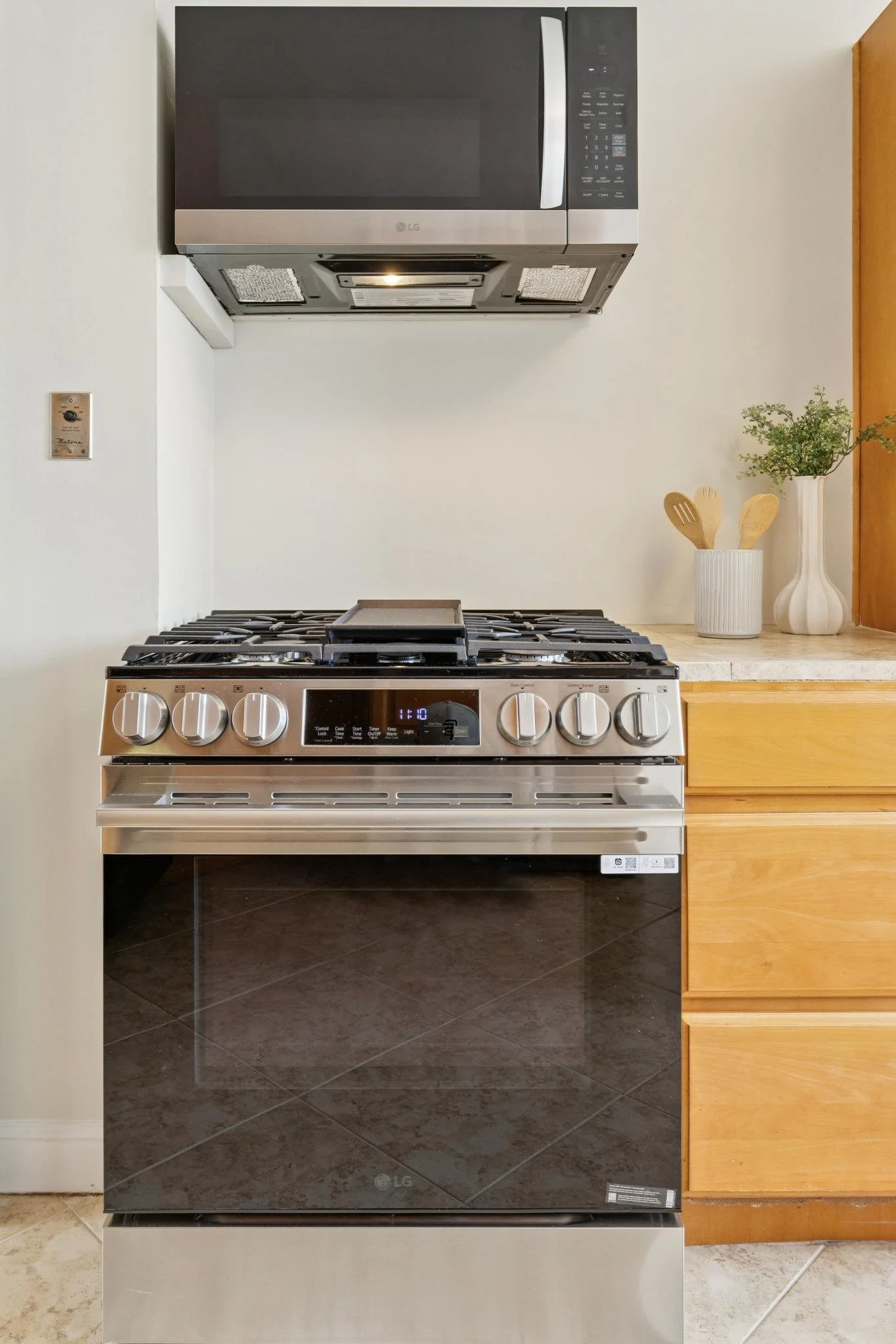 A modern kitchen with a stainless steel LG microwave mounted above a stainless steel LG gas oven. To the right, there are wooden drawers and a white vase with plants and wooden utensils. The floor has dark tiles.